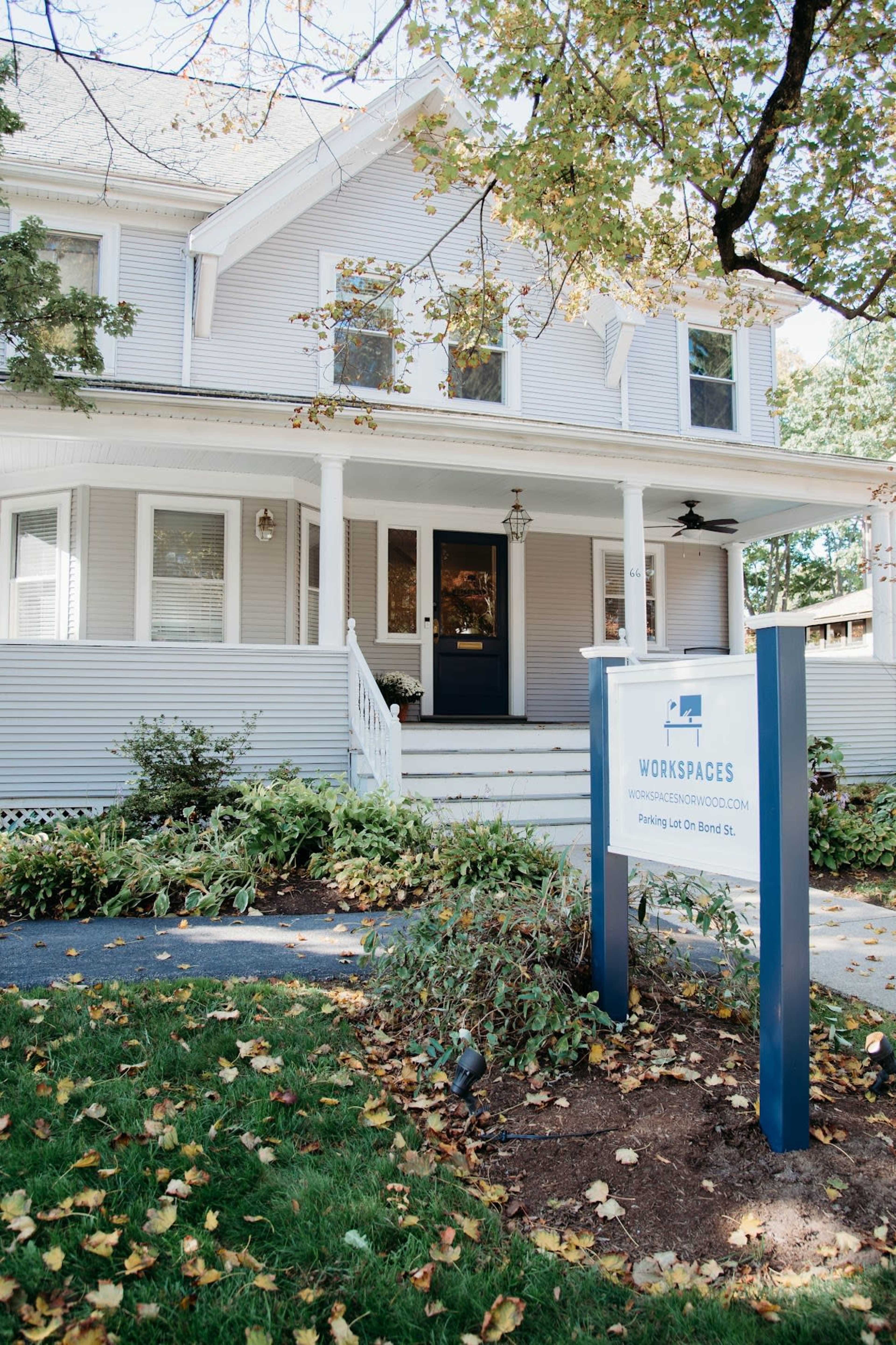 A light-colored, two-story house with a front porch and a sign advertising workspaces stands in a landscaped area with autumn leaves on the ground.