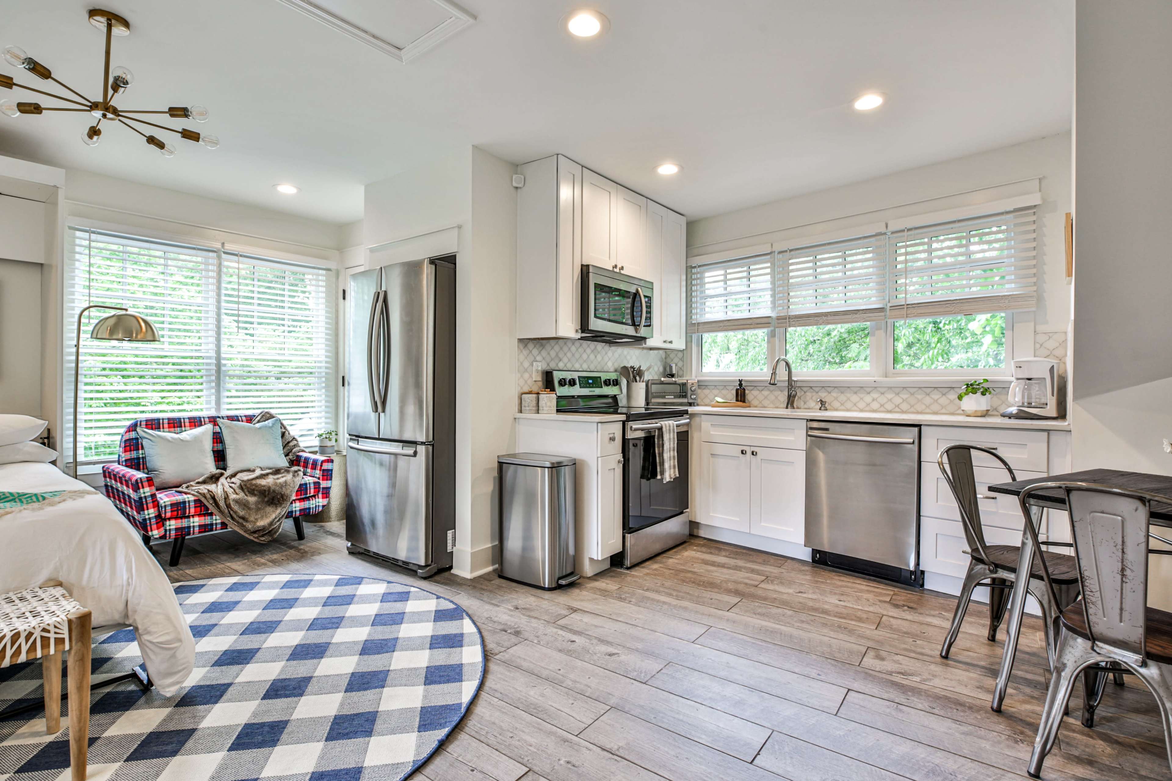 A bright, modern studio apartment featuring a small kitchen area, a dining set, and a bed beside a window with blinds.