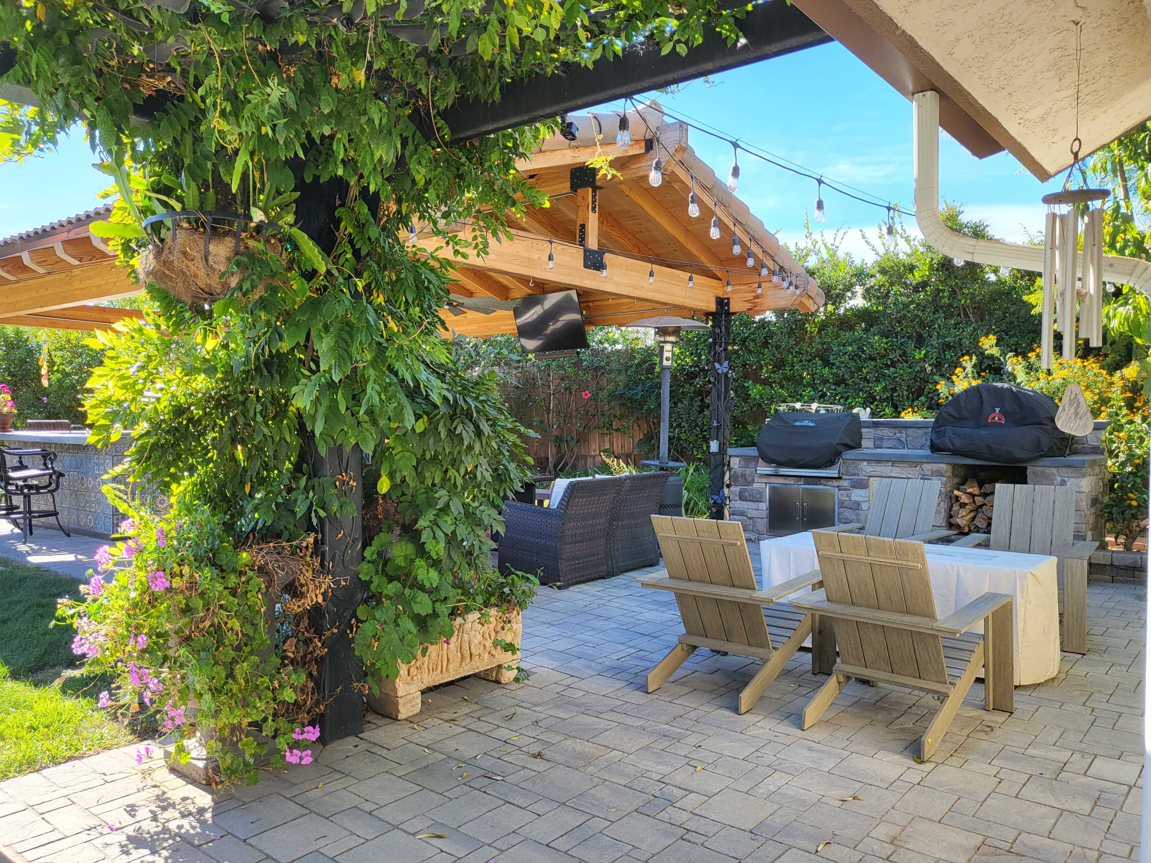 The image shows an outdoor patio area featuring a covered seating space with wooden furniture, a barbecue grill, and lush greenery surrounding the space.