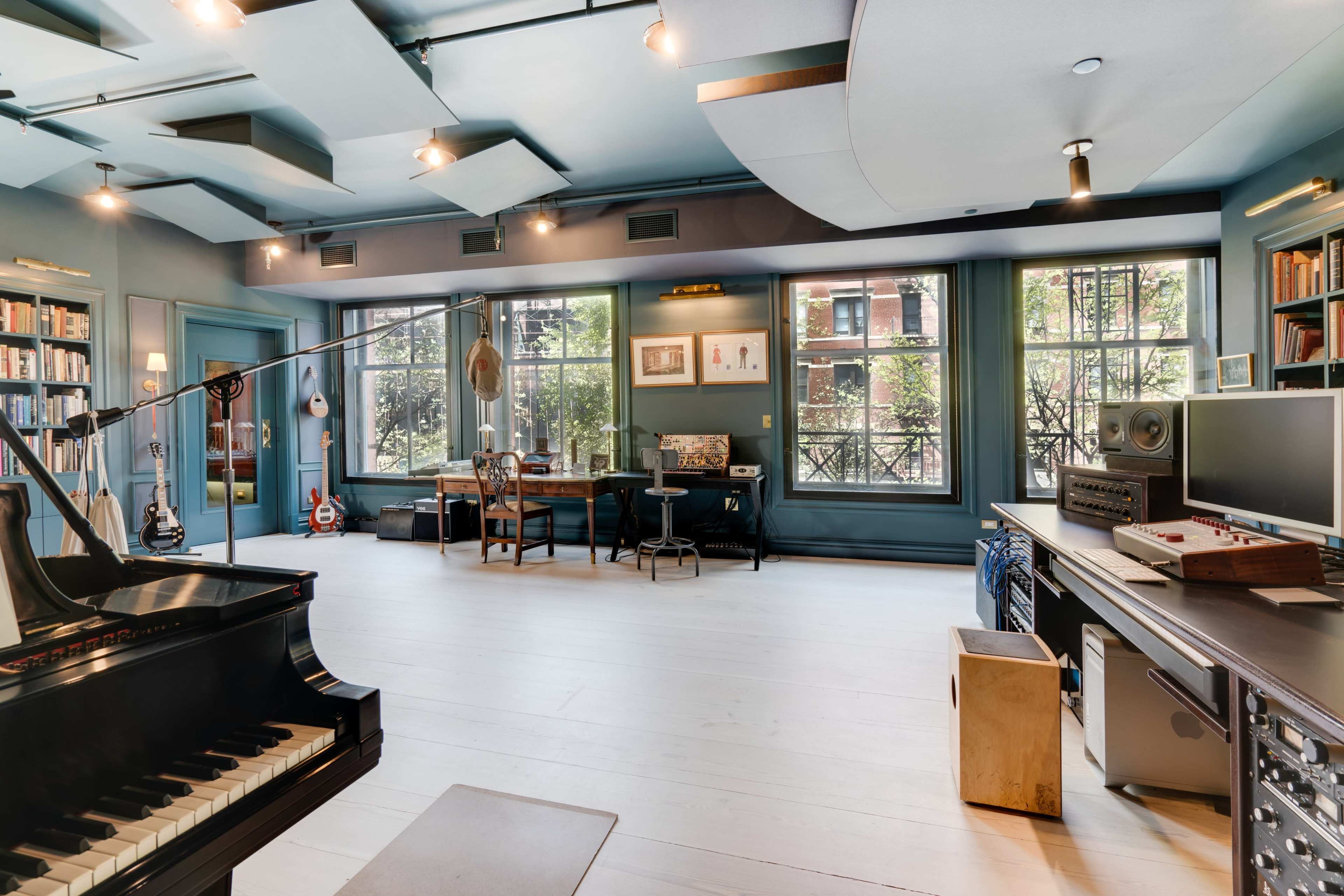 A spacious music studio features a grand piano, recording equipment, and a table surrounded by windows that overlook a garden.