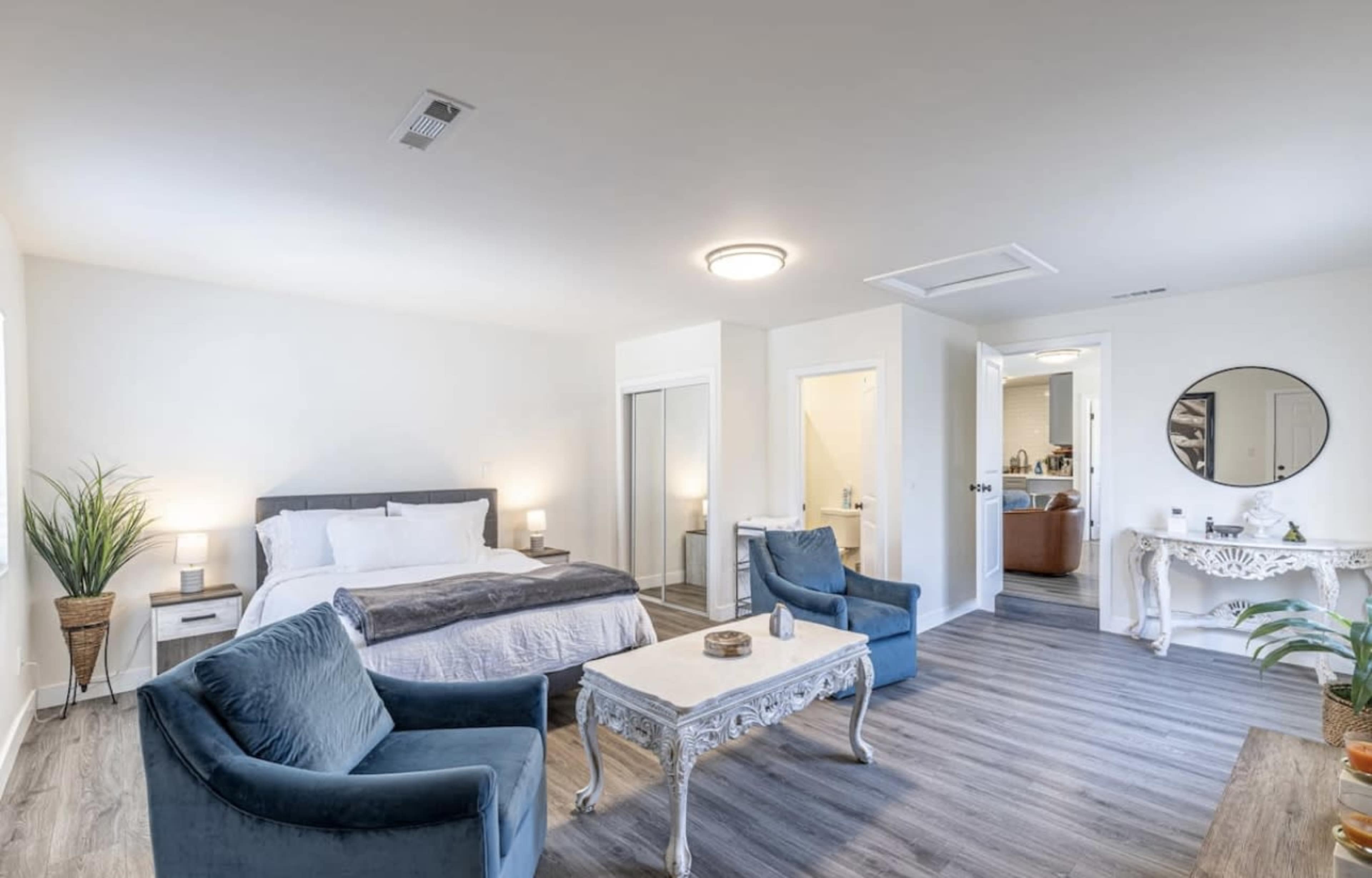 A spacious bedroom featuring a bed, two armchairs, a coffee table, and an ensuite bathroom visible in the background.