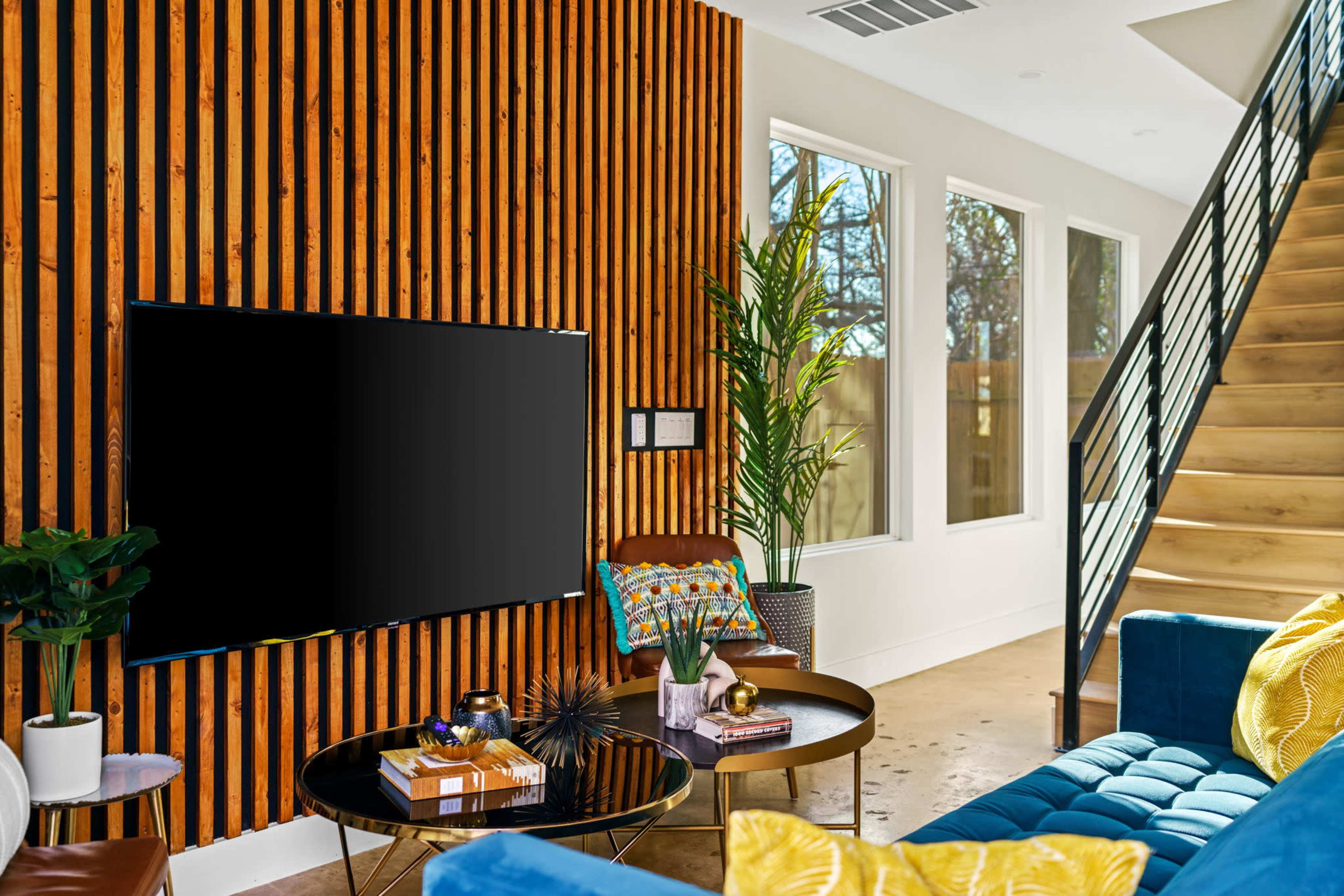 A modern living room features a large wall-mounted television, a blue sofa with yellow accents, and a wooden staircase beside a wall with vertical wooden slats.