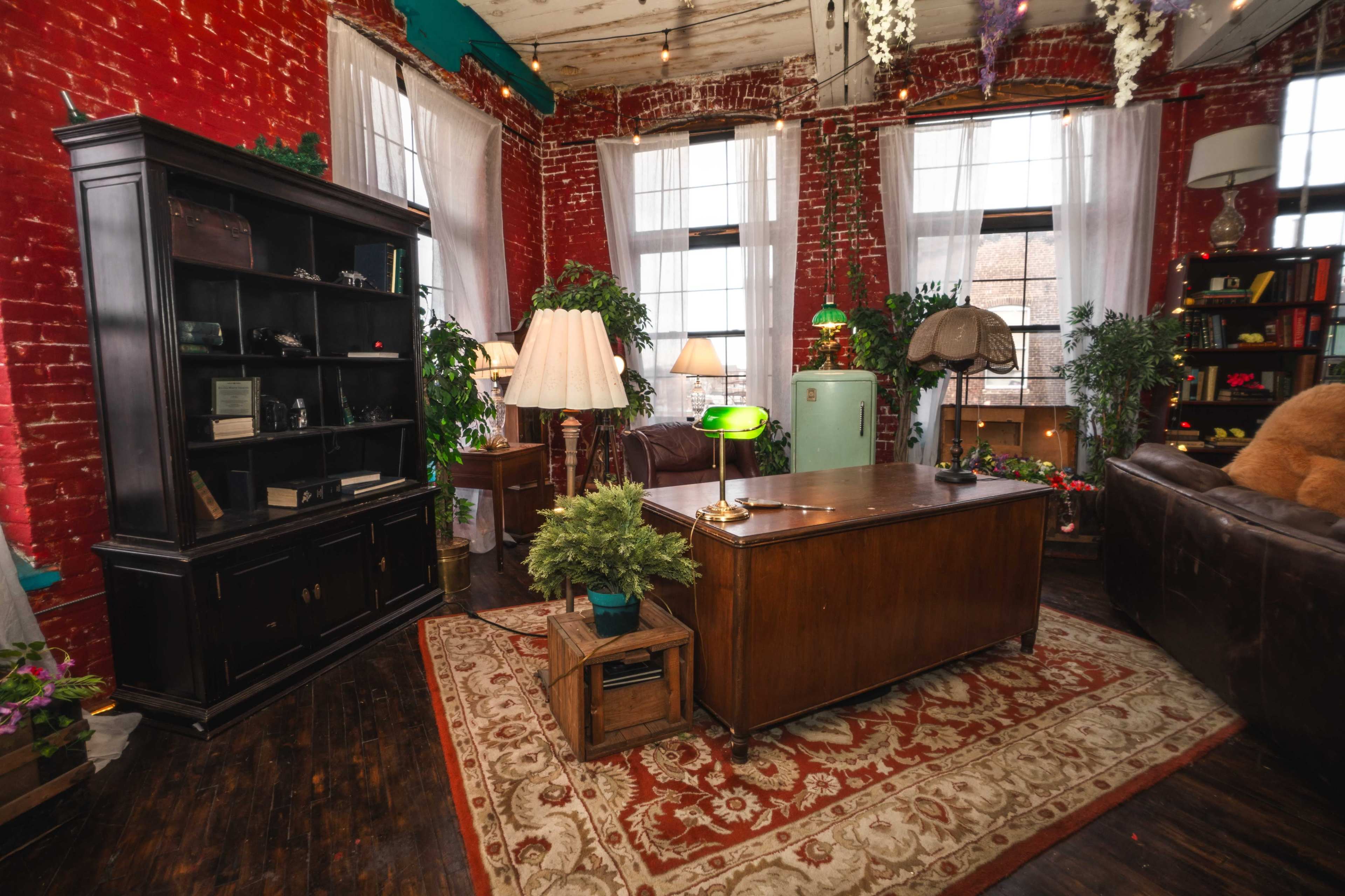 The image shows a cozy room with red brick walls, featuring a wooden desk, shelves filled with books, plants, and vintage lamps.