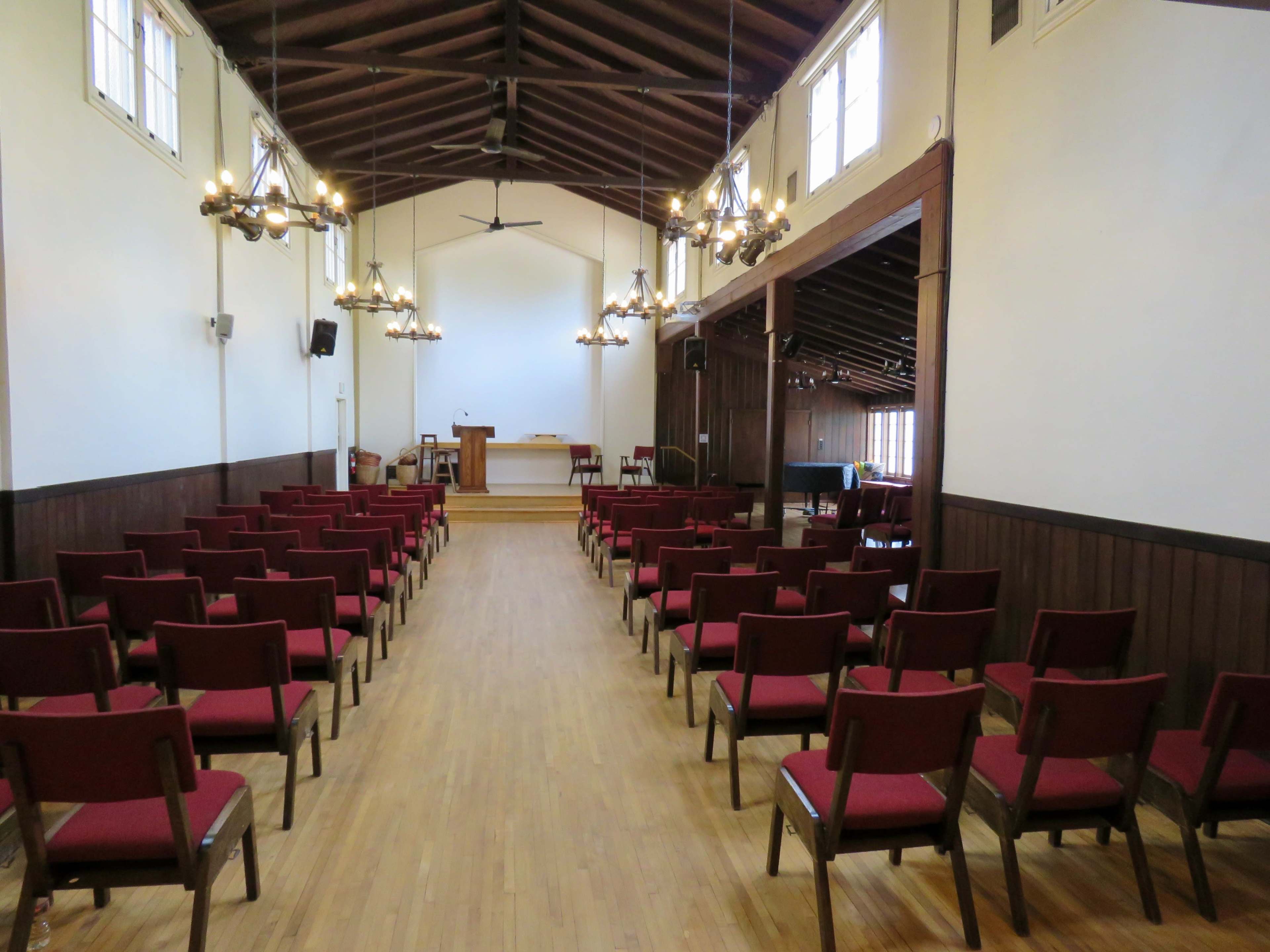 A spacious room with wooden floors, lined with rows of red chairs facing a raised platform at the front.