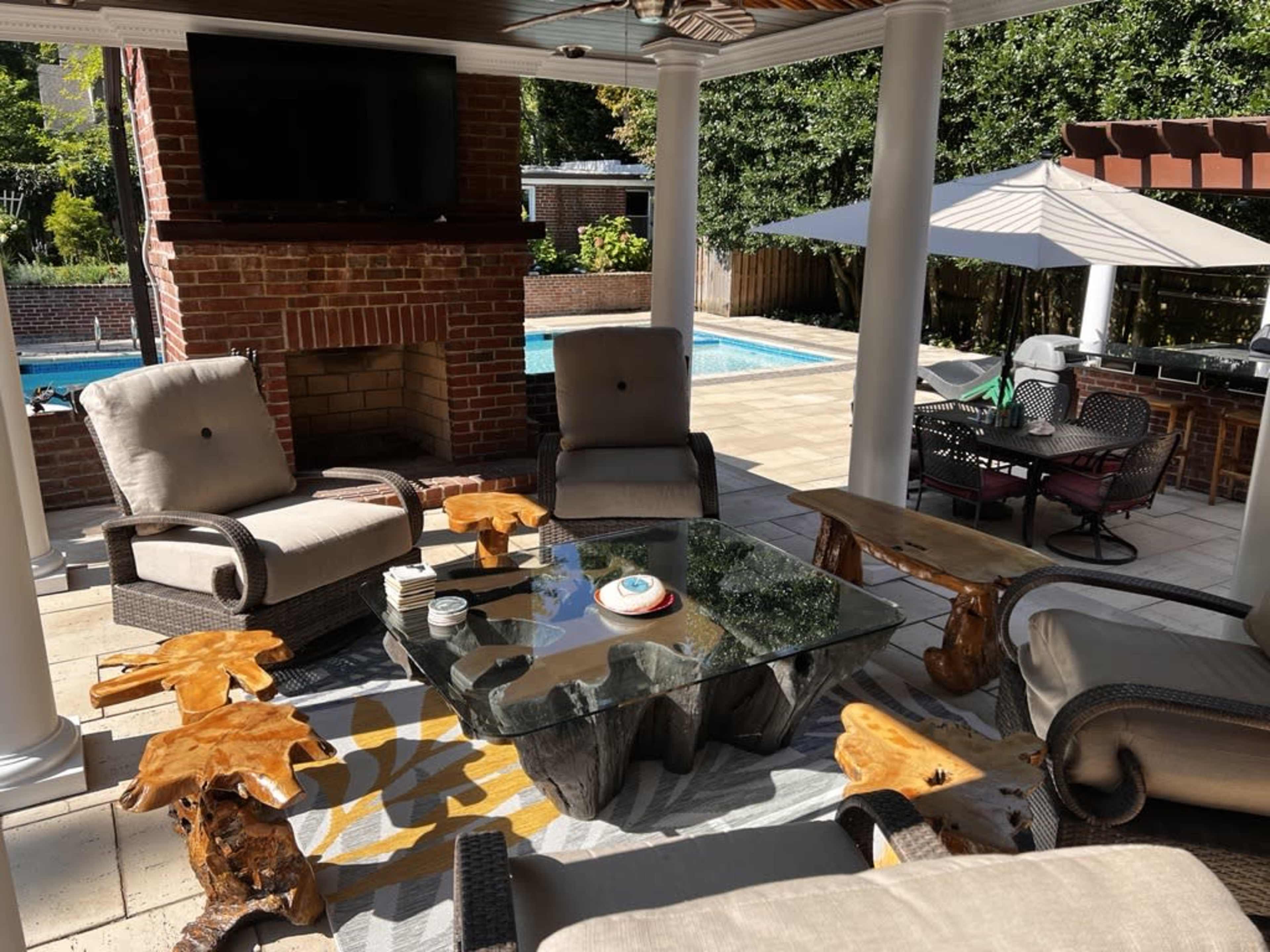 A patio area features comfortable seating, a glass coffee table, and a view of a swimming pool surrounded by landscaping.