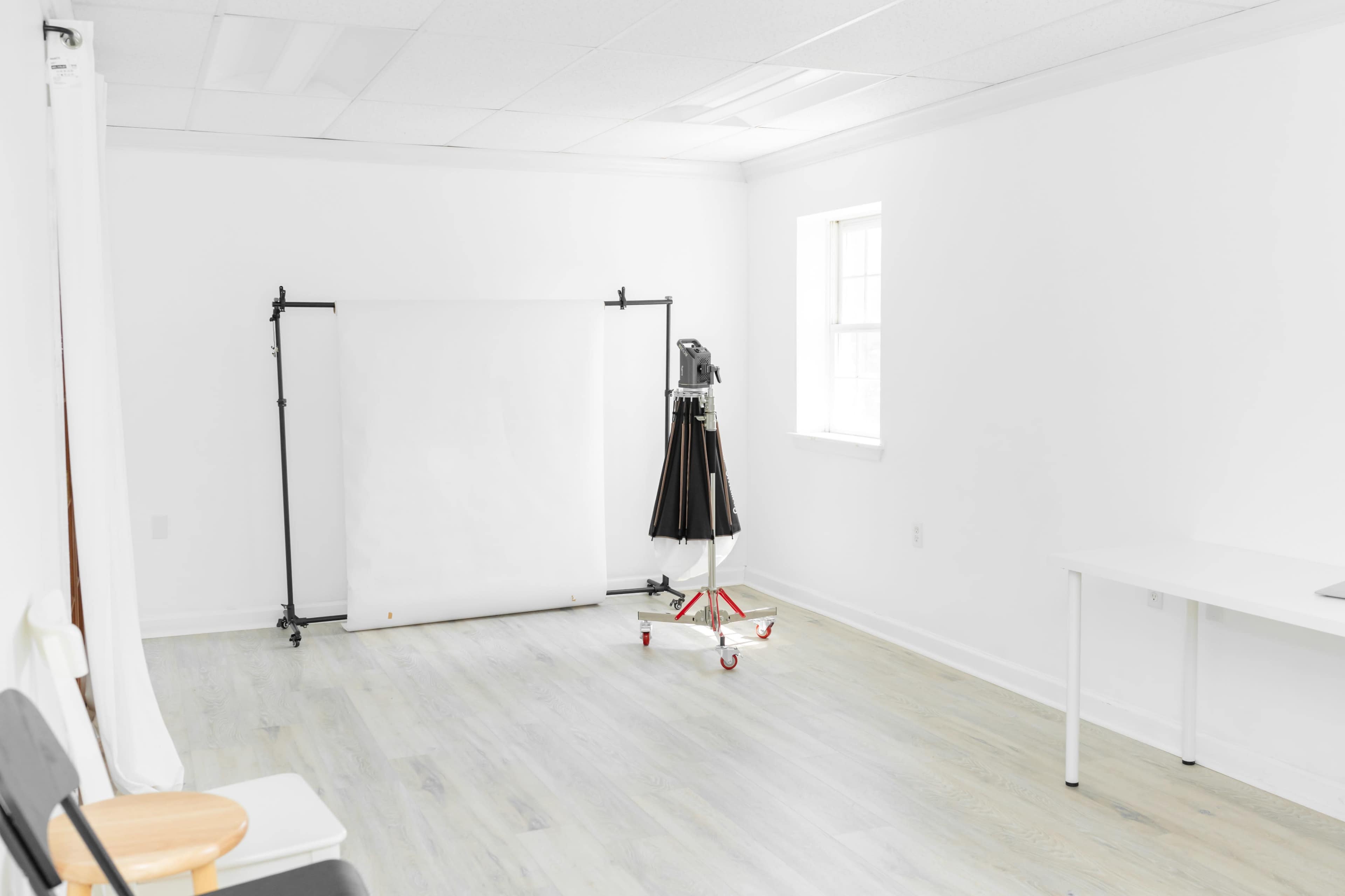 A minimalistic, white studio space featuring a blank backdrop on a stand, a garment rack, and a table.