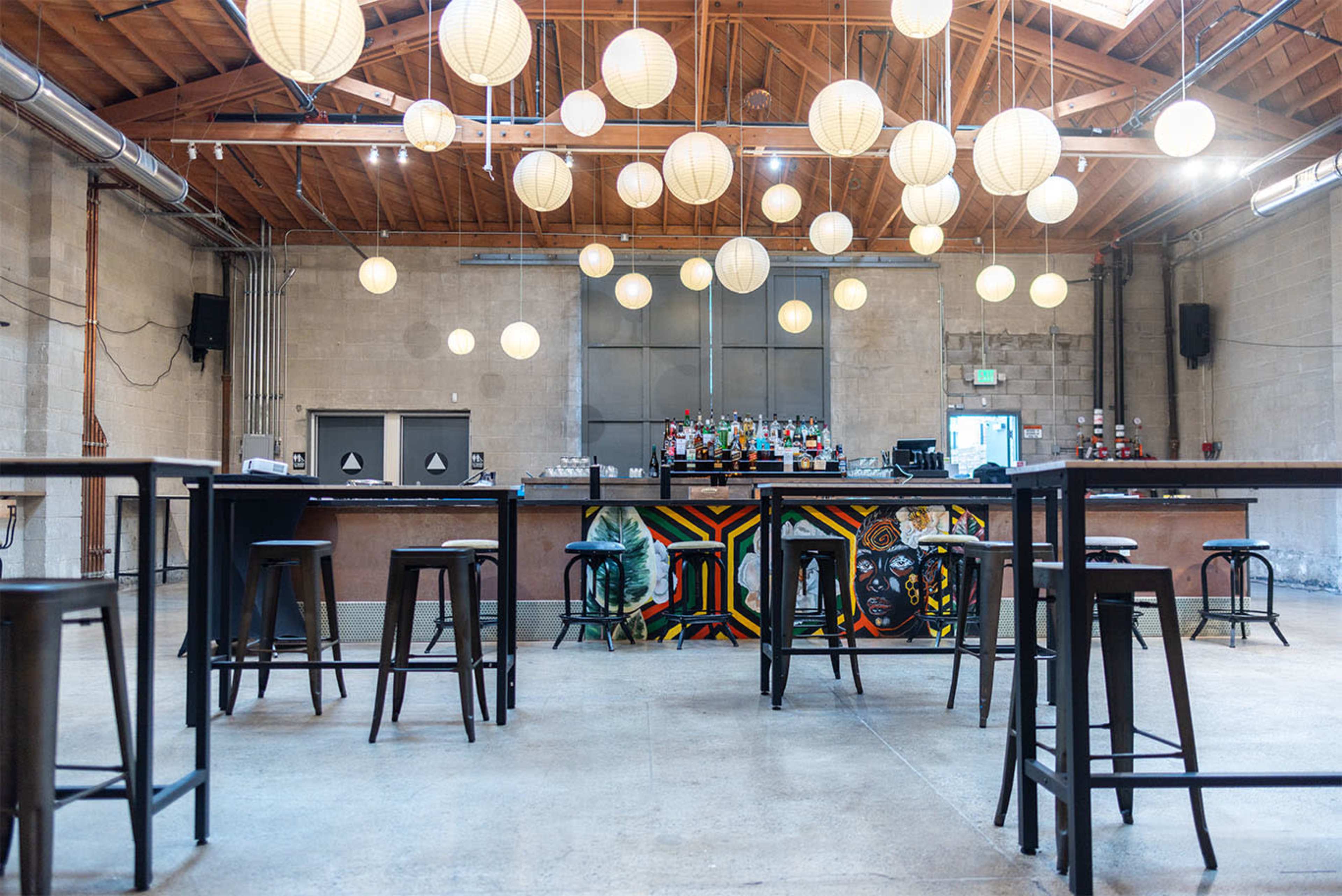 The image shows a spacious bar area with high tables and stools, illuminated by hanging paper lanterns, against a backdrop of a well-stocked bar and textured walls.
