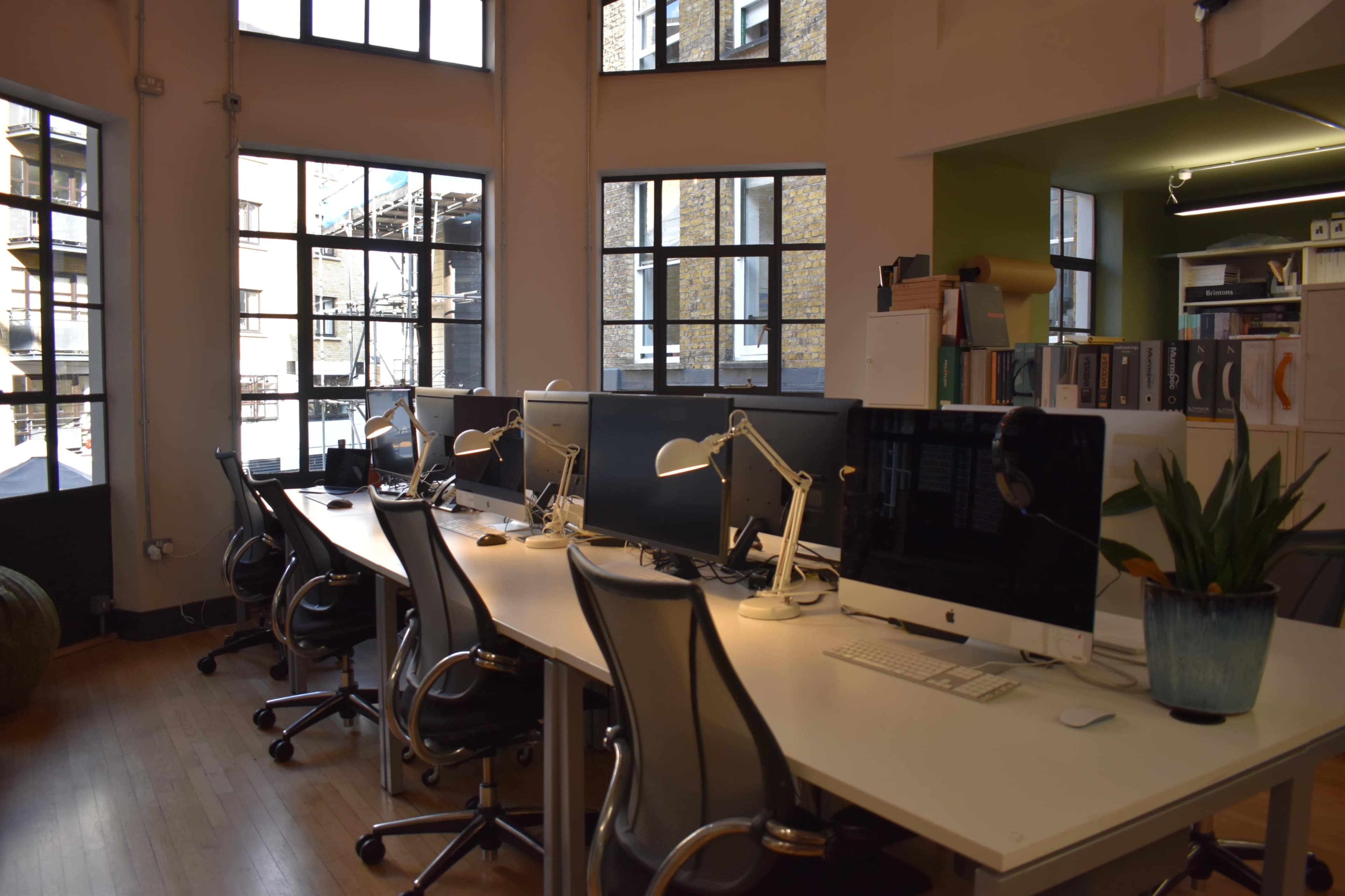 The image shows a modern office space with multiple workstations featuring desk computers and ergonomic chairs, illuminated by natural light through large windows.