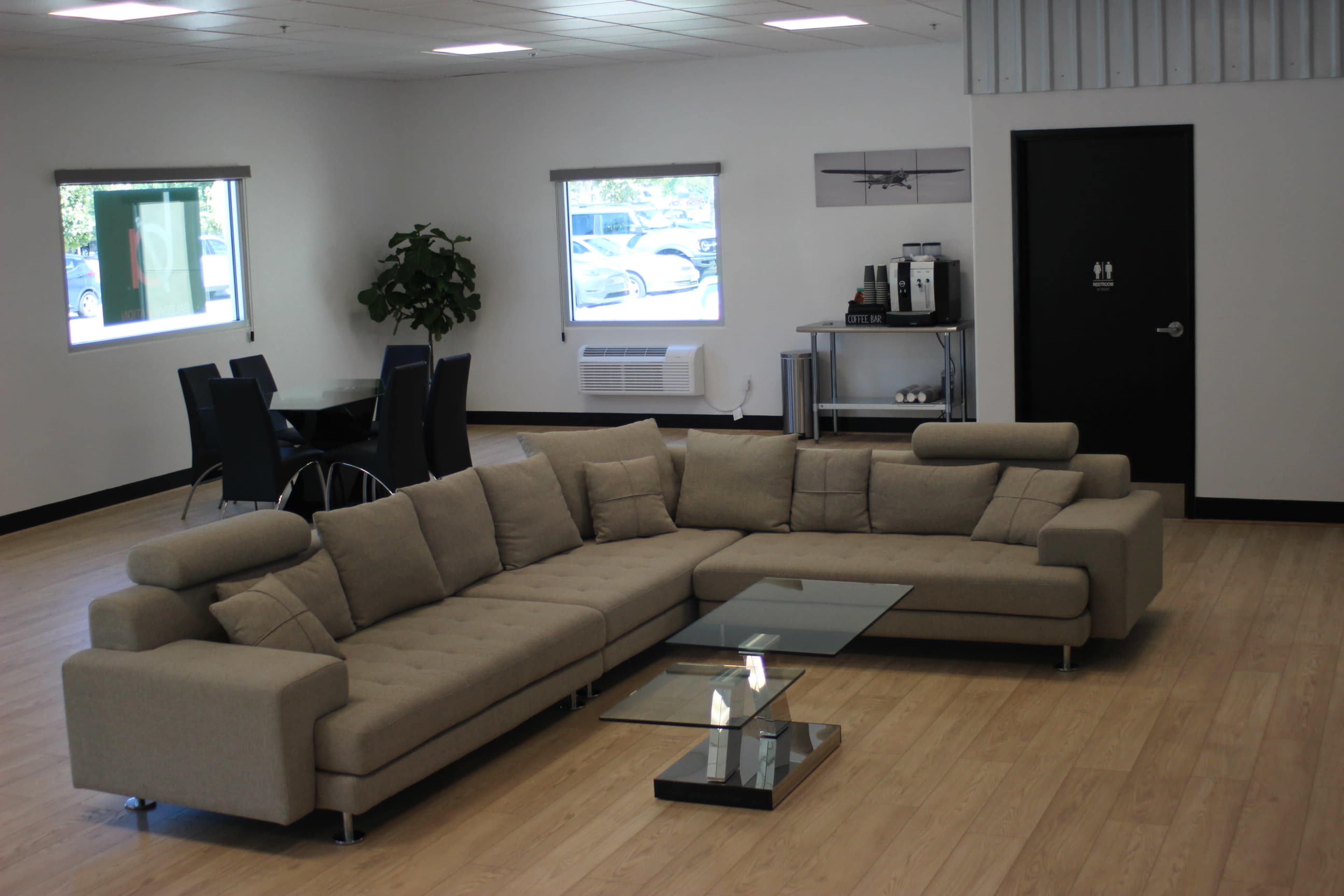 A modern lounge area featuring a large sectional sofa with a glass coffee table, surrounded by a dining table and chairs, and a kitchenette in a well-lit room with light wood flooring.