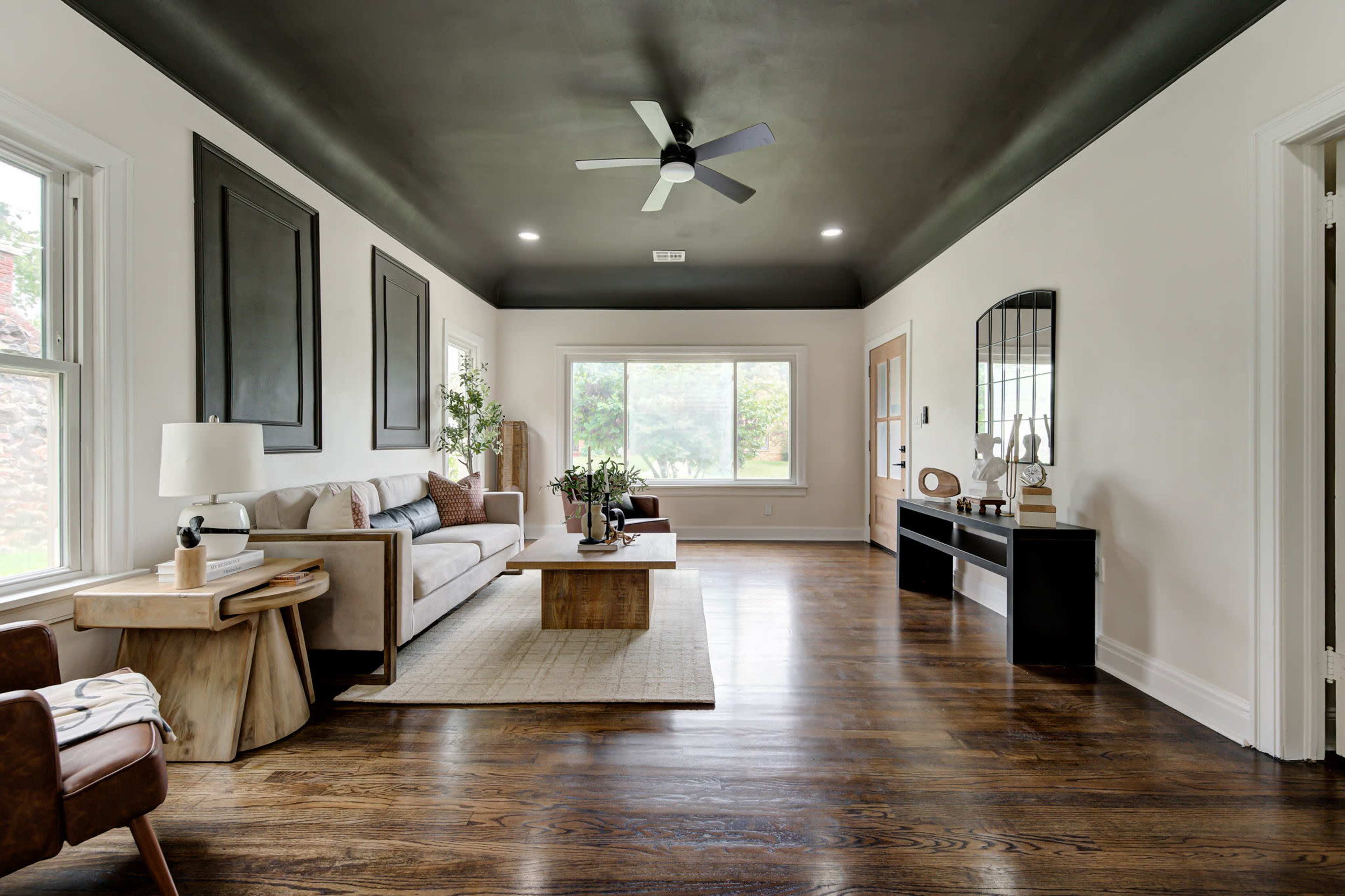 The living room features a light-colored sofa, a decorative coffee table, a dark ceiling, and large windows allowing natural light to enter.