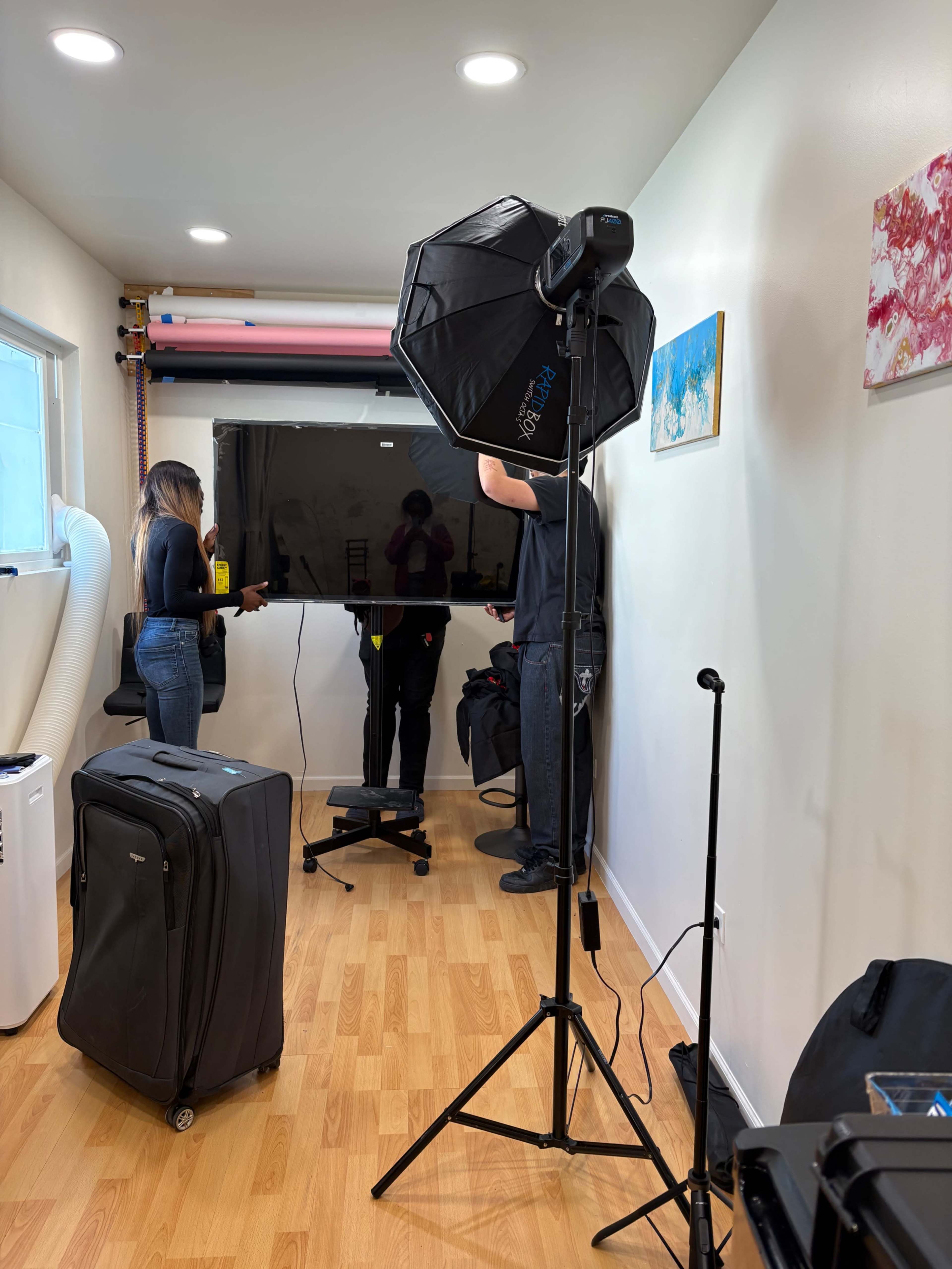 A photography studio features a camera setup with a softbox, a television on a stand, and a person preparing for a shoot.