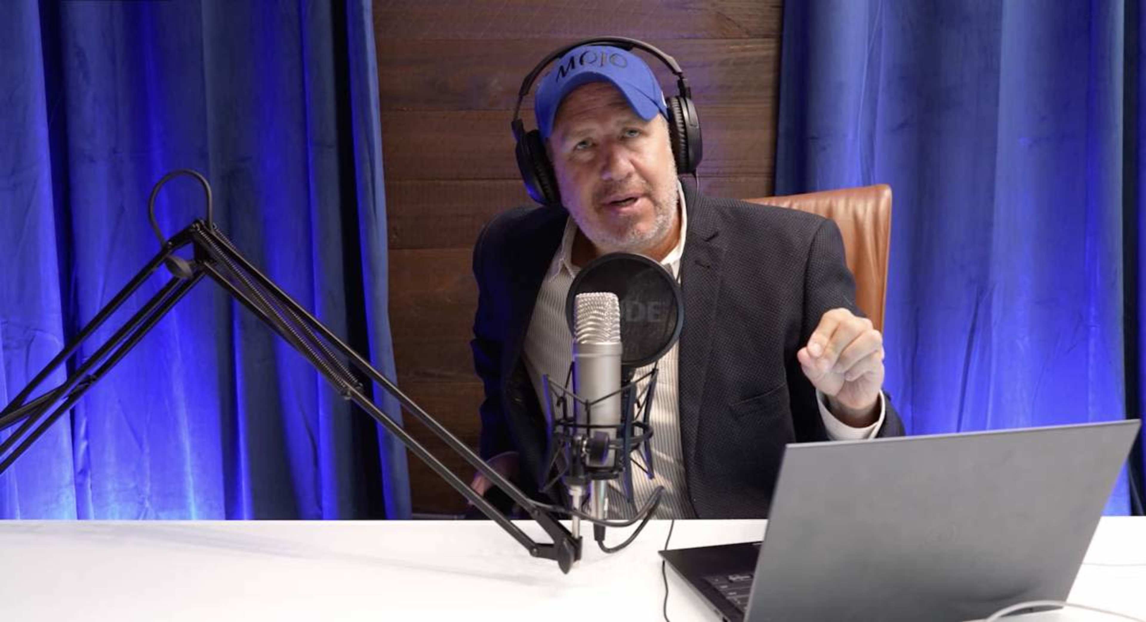 A man wearing a blue cap and headphones speaks into a microphone while seated at a desk with a laptop.