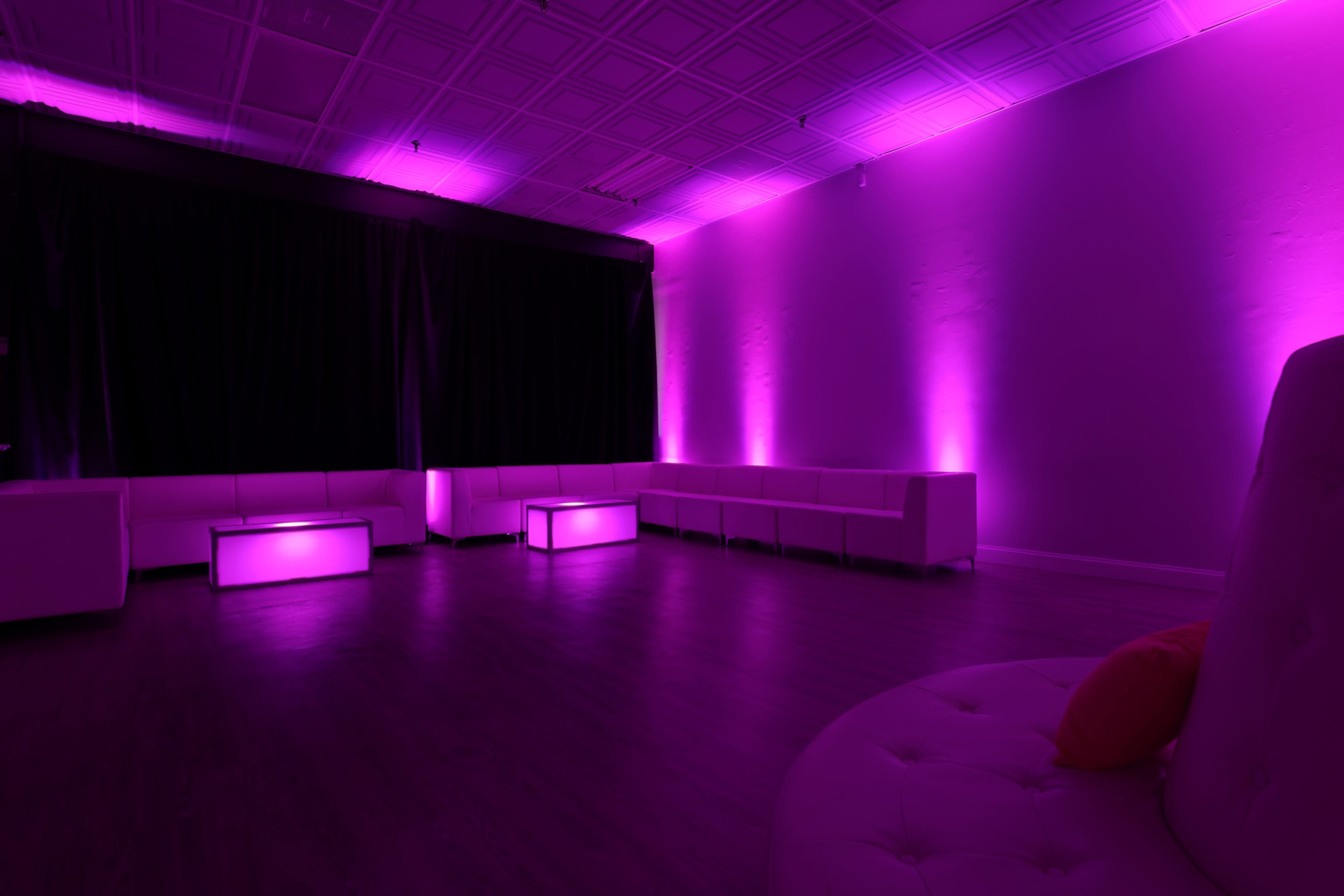 The image shows a modern lounge space with purple lighting, featuring white seating and illuminated coffee tables.