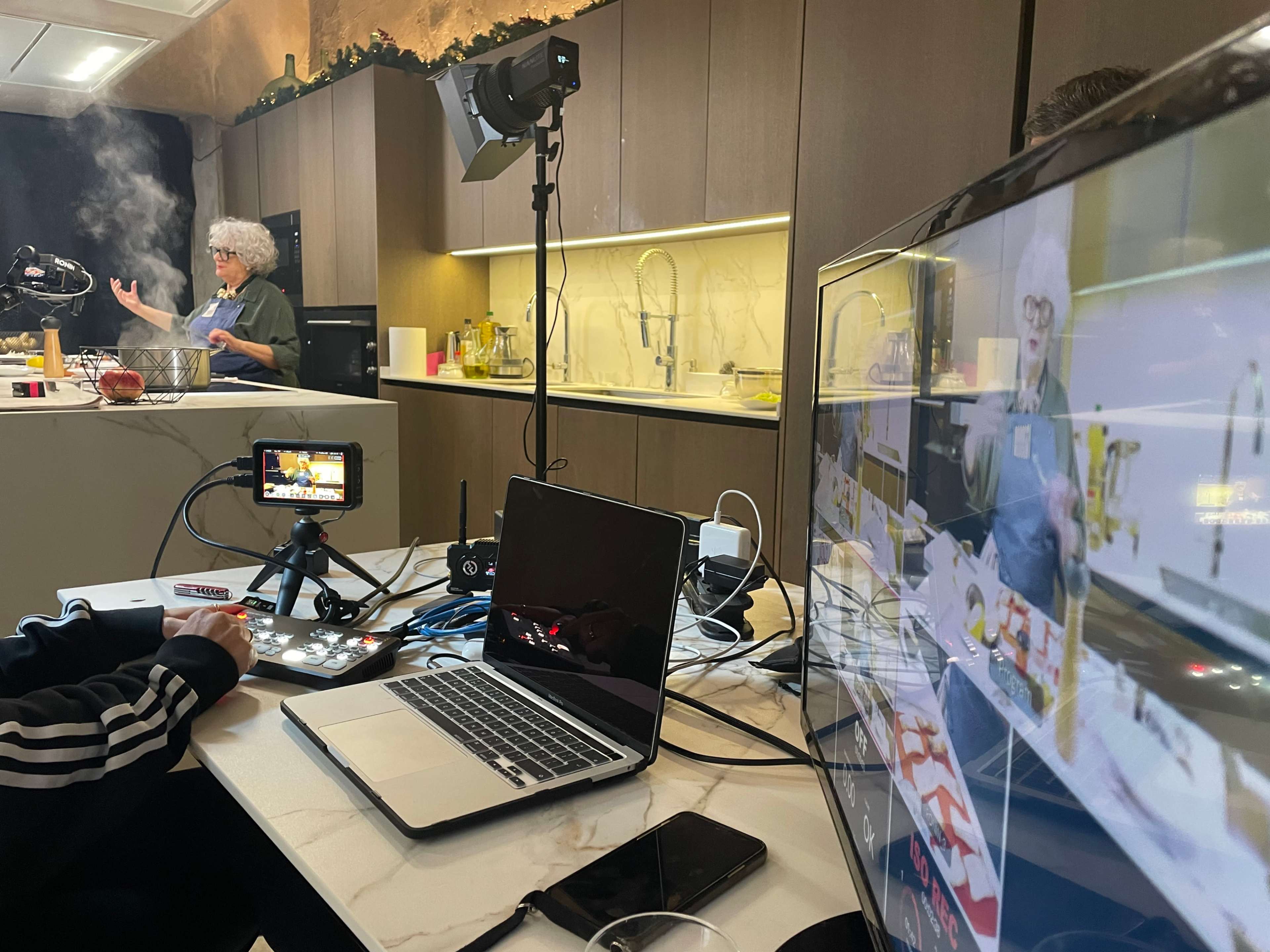 A kitchen is set up for a cooking demonstration, featuring a chef preparing food while cameras and equipment capture the event.
