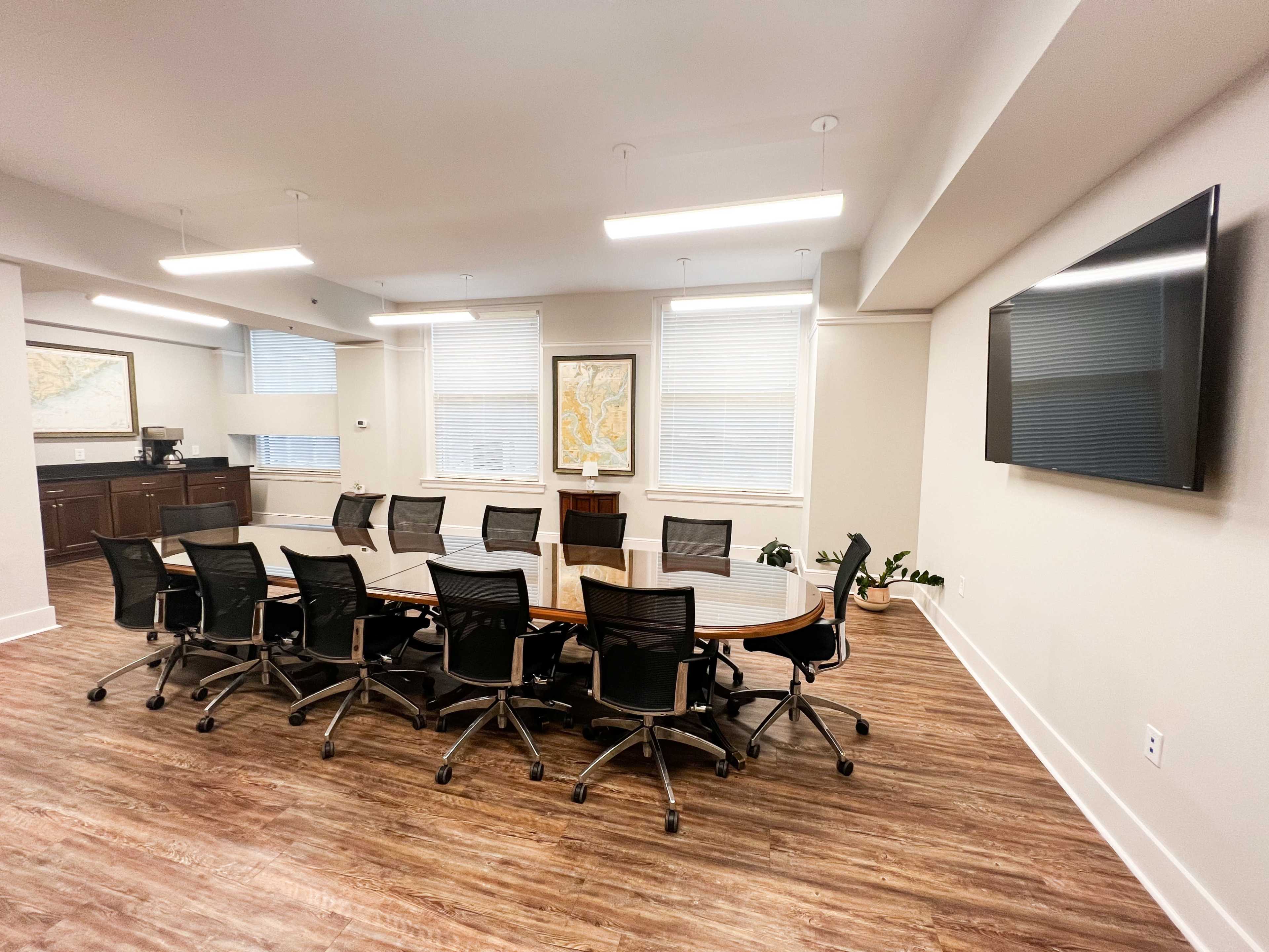 A modern conference room features a long wooden table surrounded by black office chairs, with large windows and a mounted television on the wall.