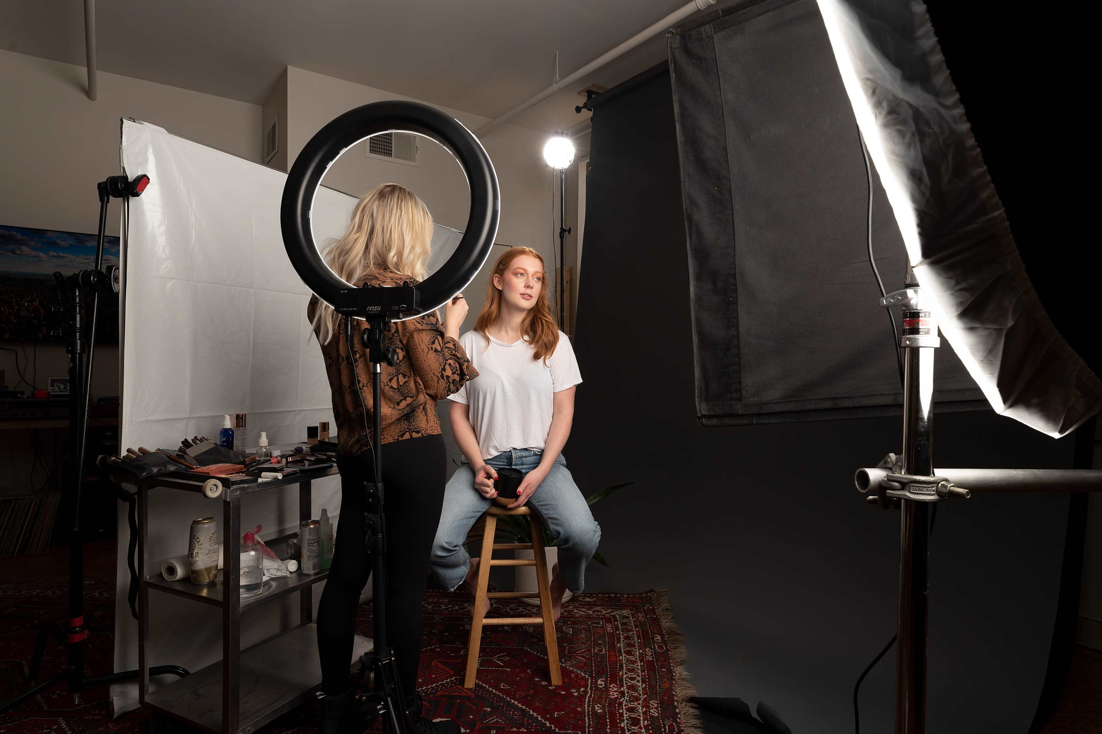 A makeup artist applies makeup to a model seated on a stool in a studio setting, surrounded by photography equipment and lighting.