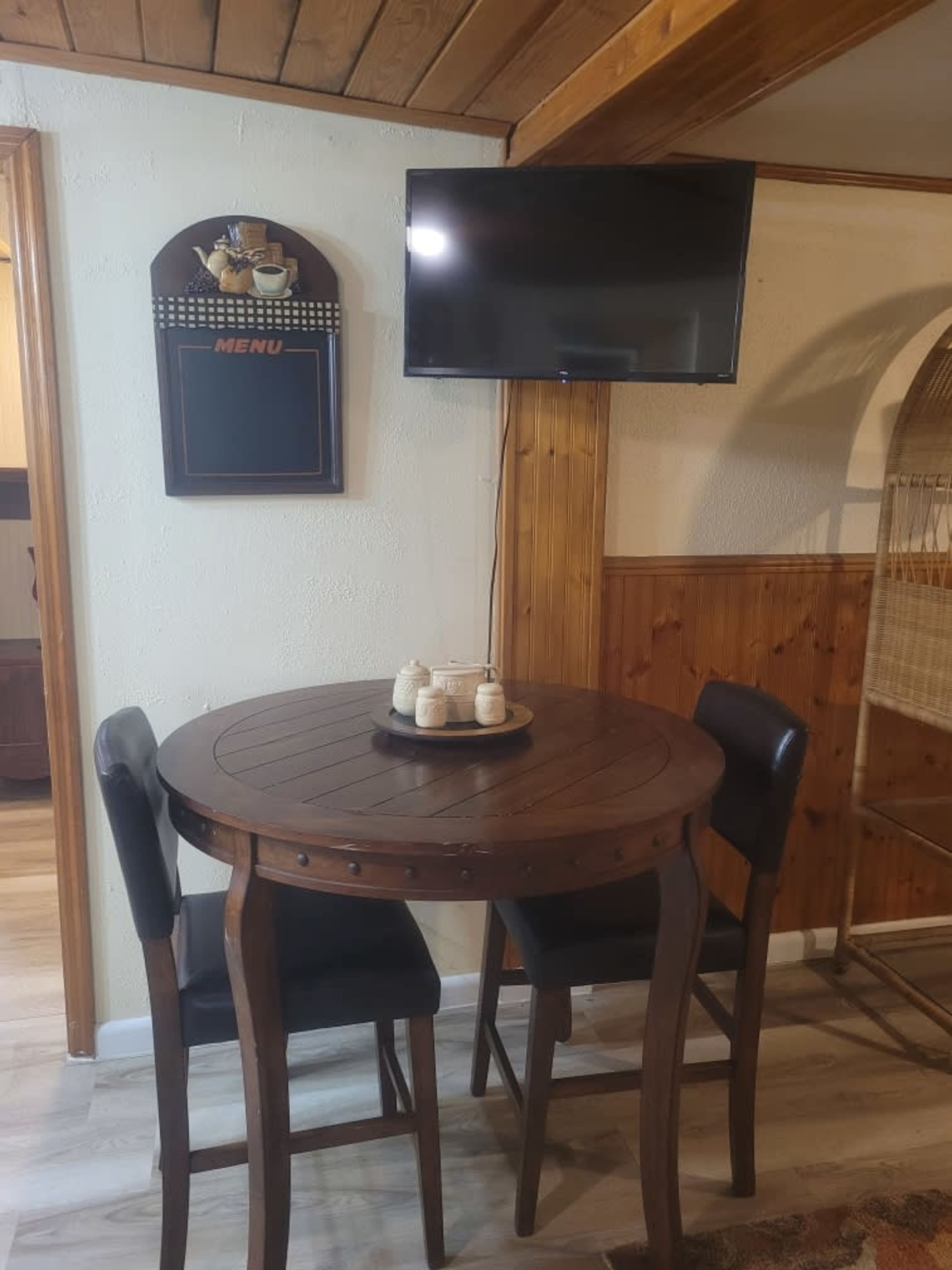 A small wooden round table with three black chairs is set against a wall with a mounted TV and a menu display.