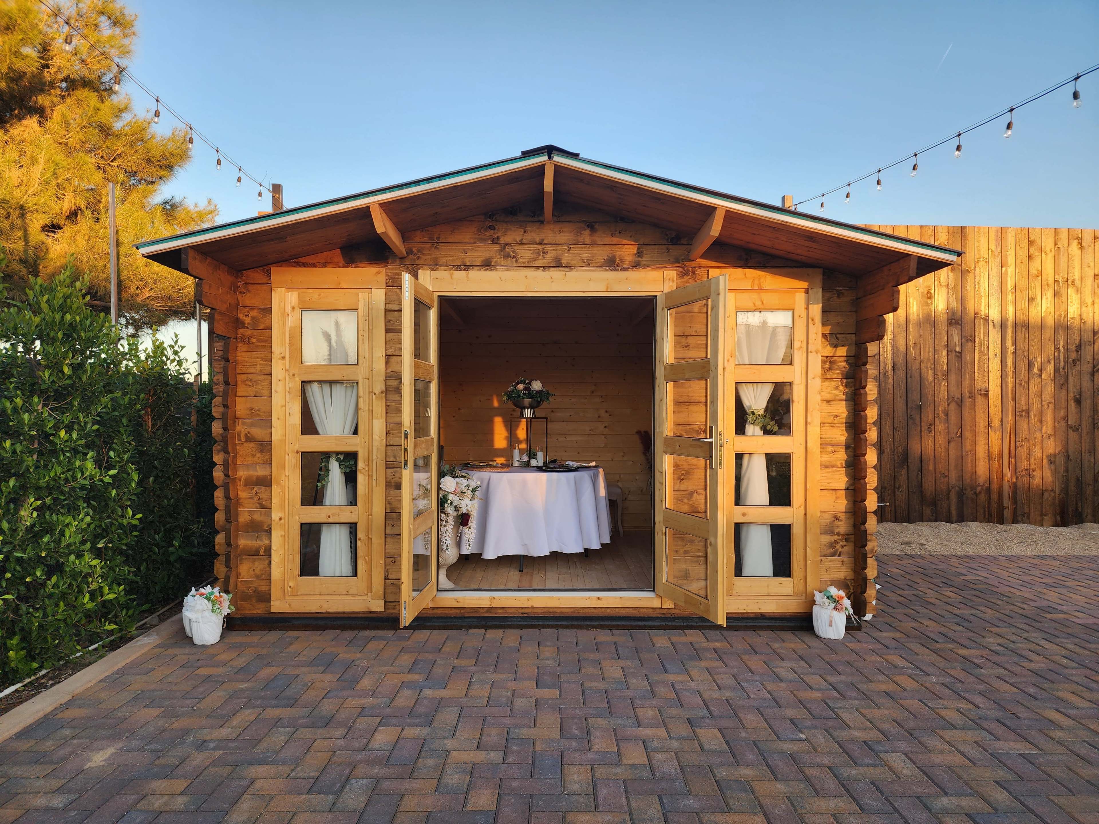 A wooden cabin with double doors is open to reveal a dining setup inside, surrounded by potted plants and a paved pathway.