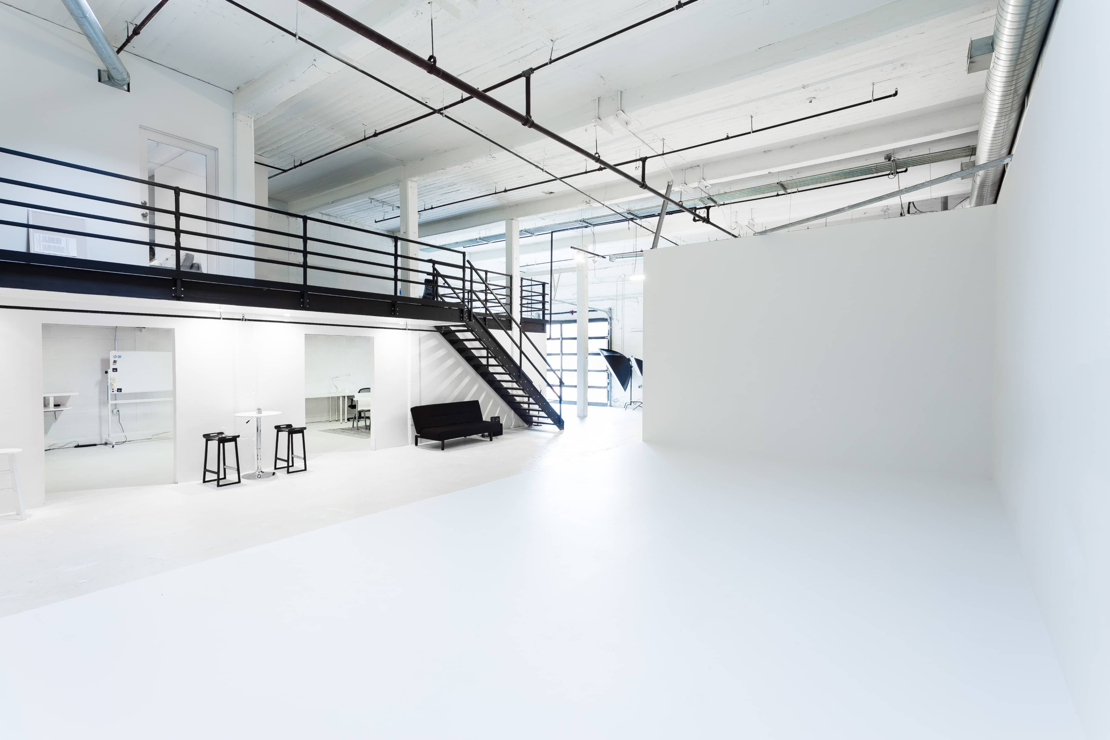 A spacious, minimalistic studio interior with white walls, a staircase leading to a mezzanine, and a black sofa placed against one side.