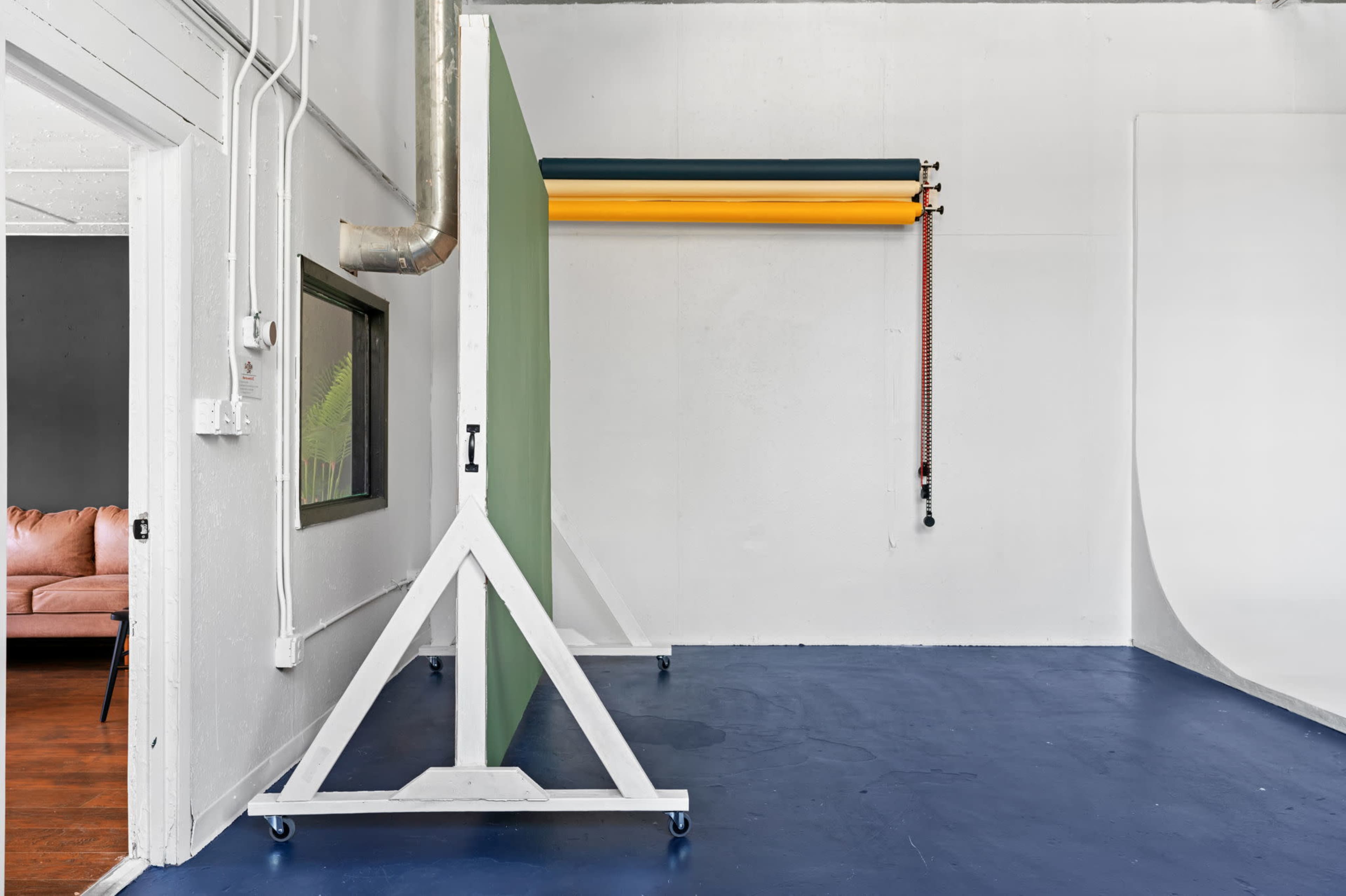 A photo studio with a green backdrop on a stand, yellow and green roll-up backdrops mounted on a wall, and a wooden frame indicating a window with a couch in the adjacent room.