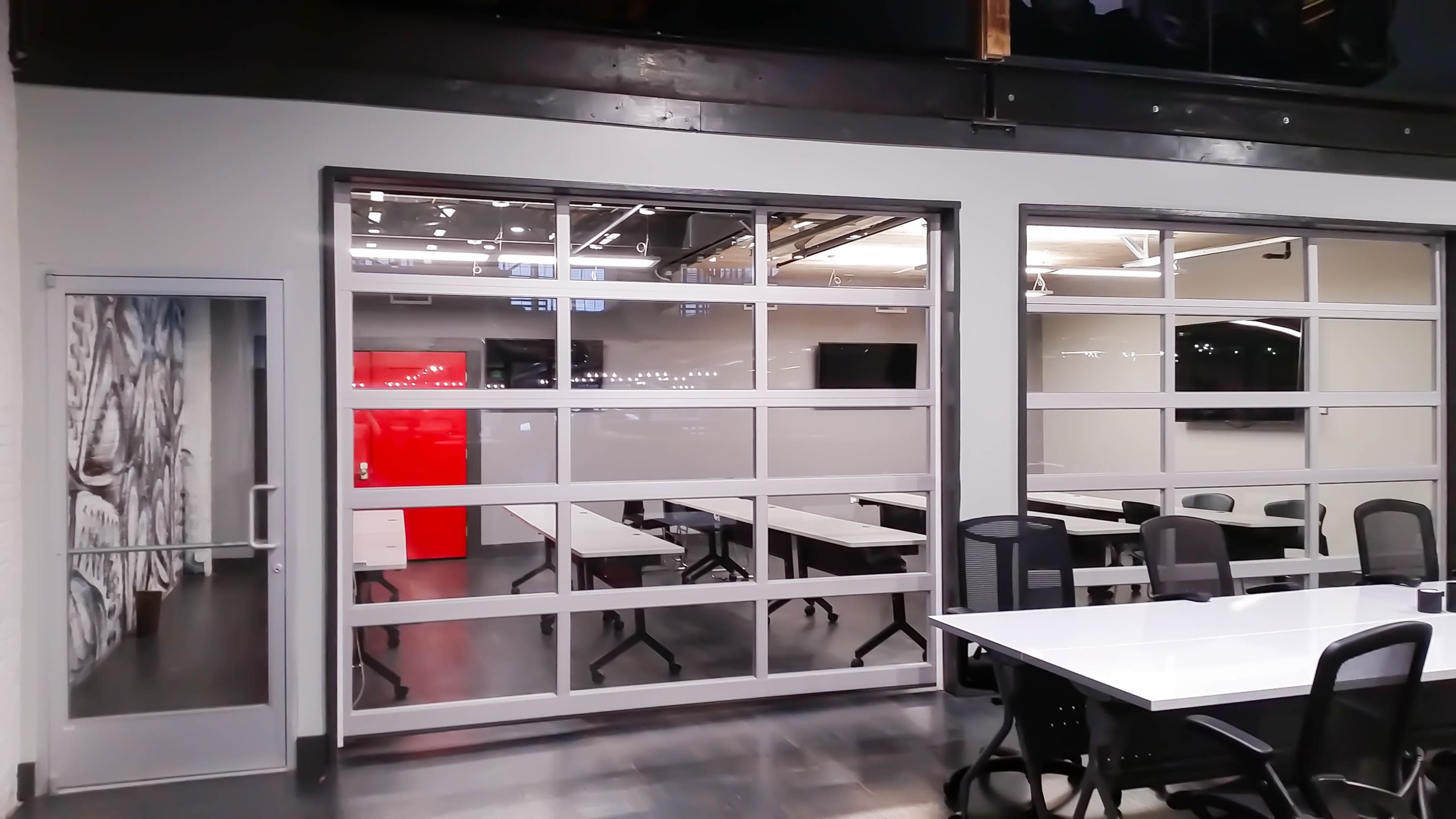 The image shows a modern office space with glass walls separating a meeting room from the main area, featuring white tables and black chairs.