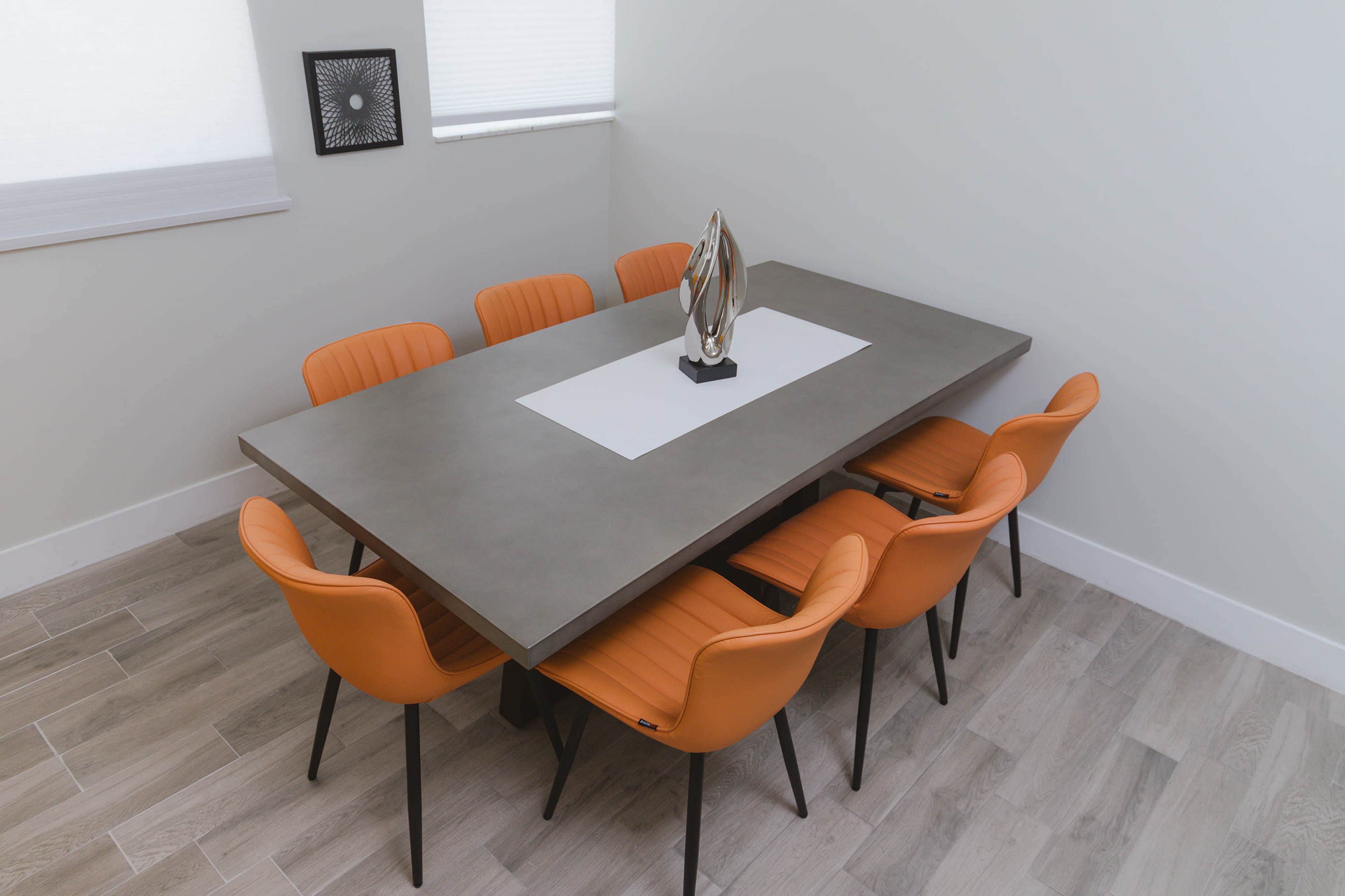 A modern dining table with a concrete top and eight orange chairs is arranged in a bright, minimalistic room.