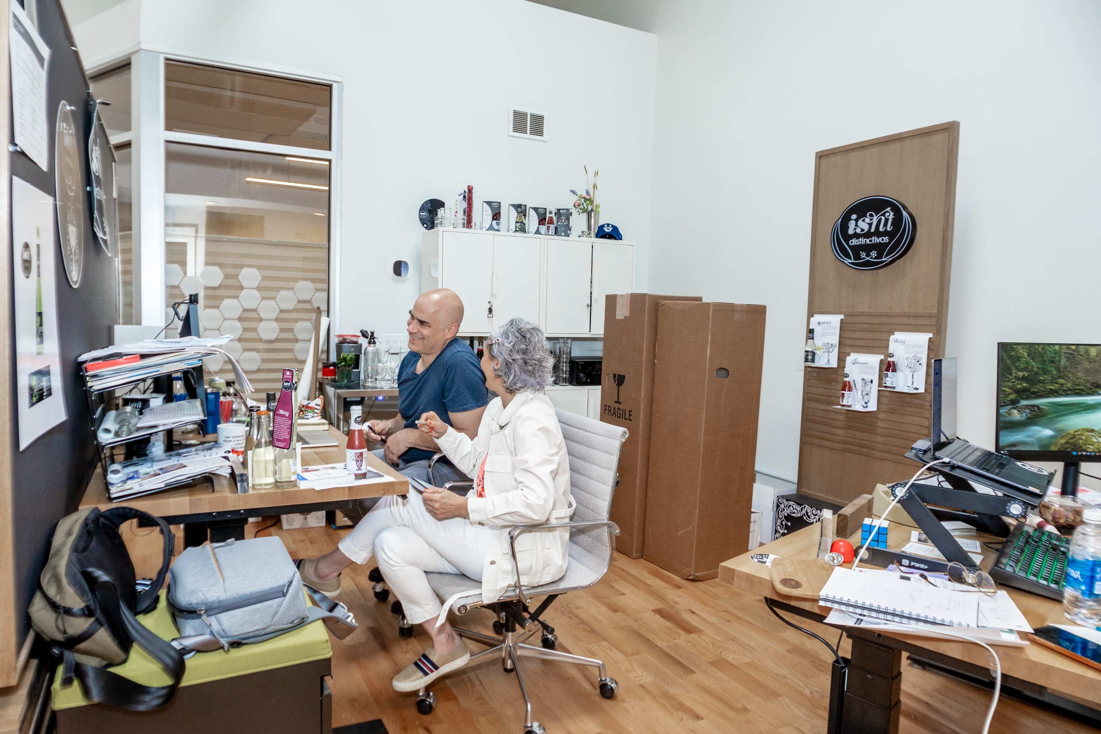A man and woman sit at a cluttered desk in an office, surrounded by boxes and various office supplies.