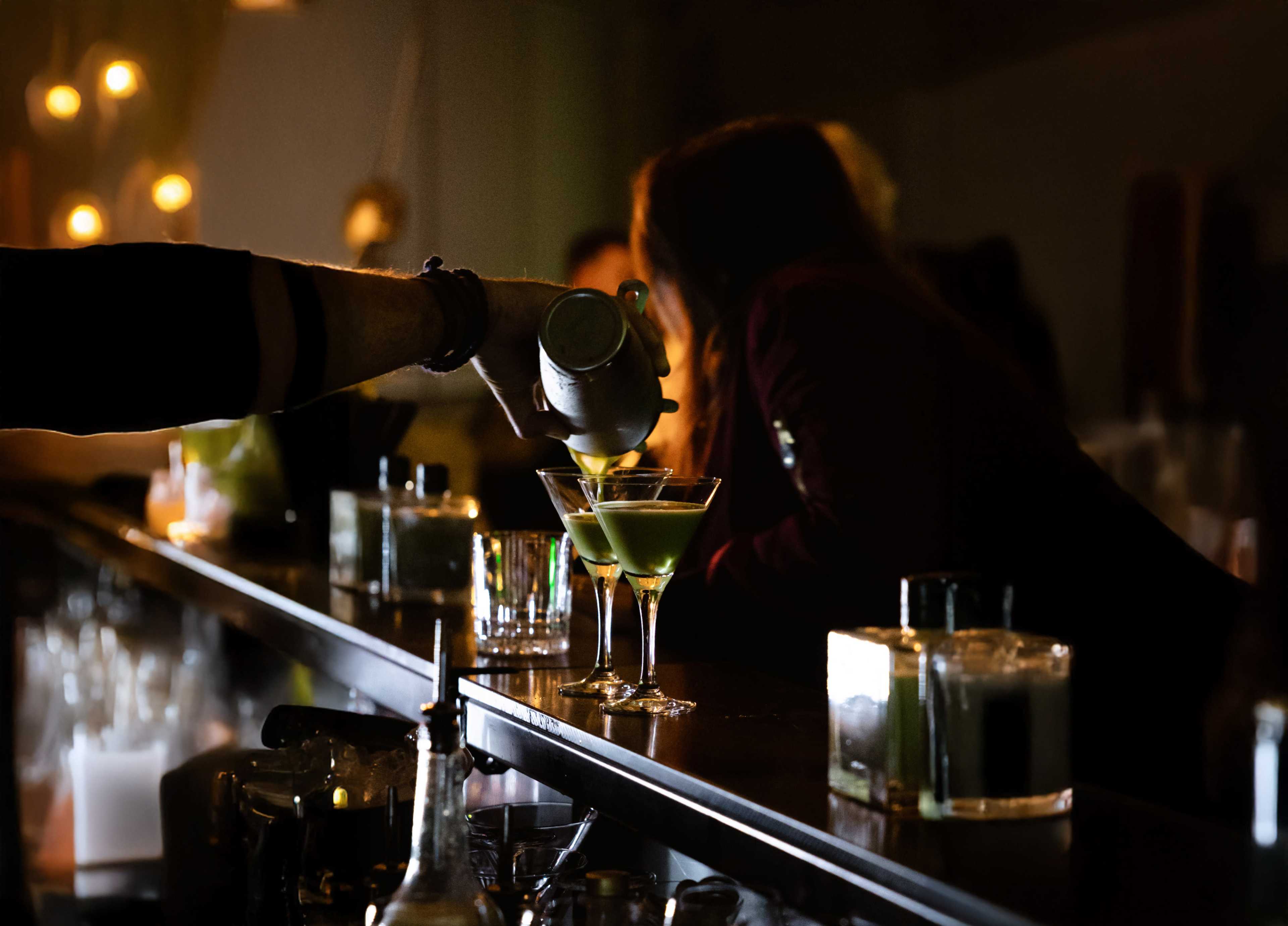 A bartender pours a cocktail into a glass at a dimly lit bar.