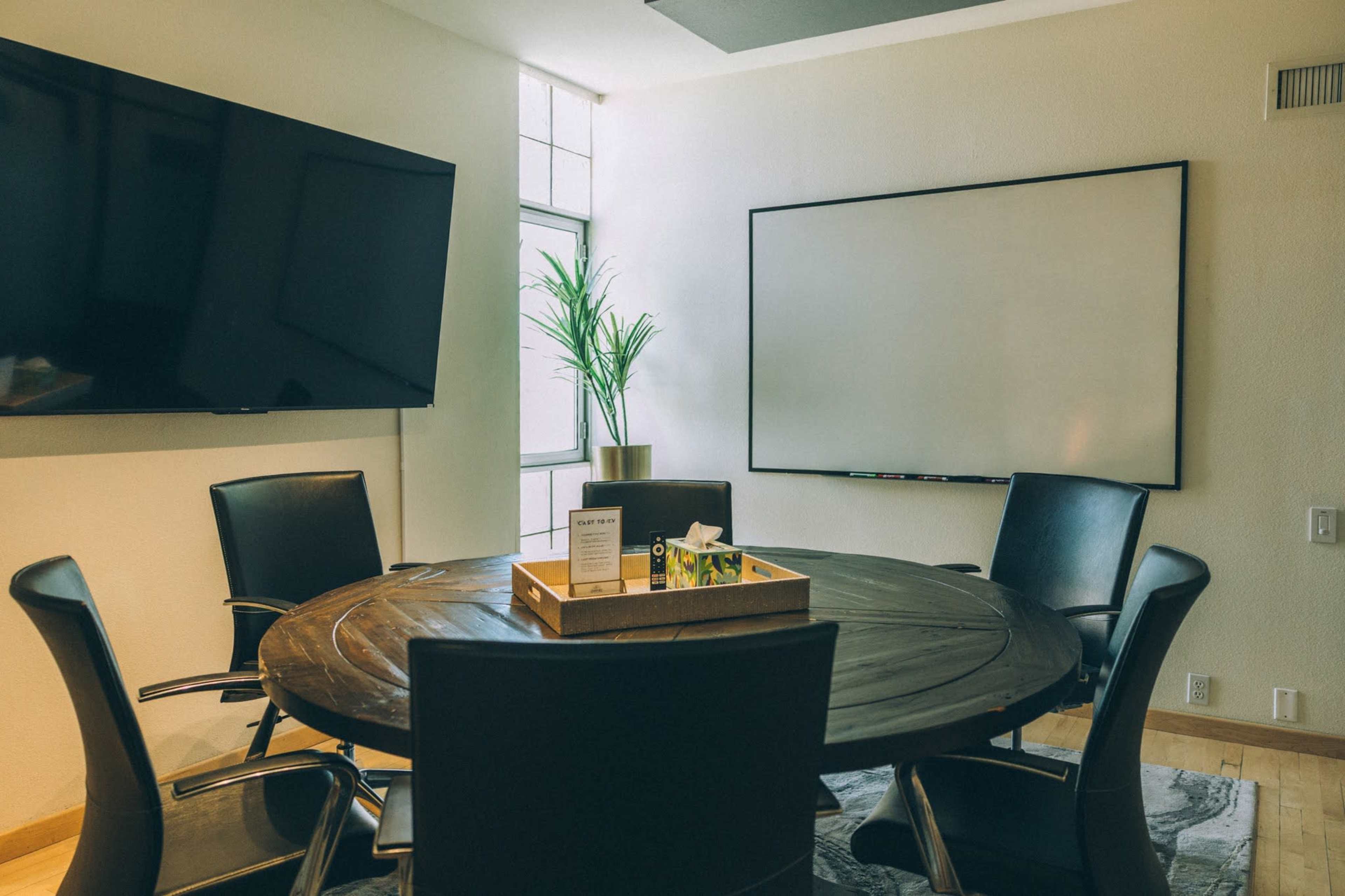 The image shows a conference room featuring a round wooden table surrounded by black chairs, with a large screen on the wall and a whiteboard nearby.