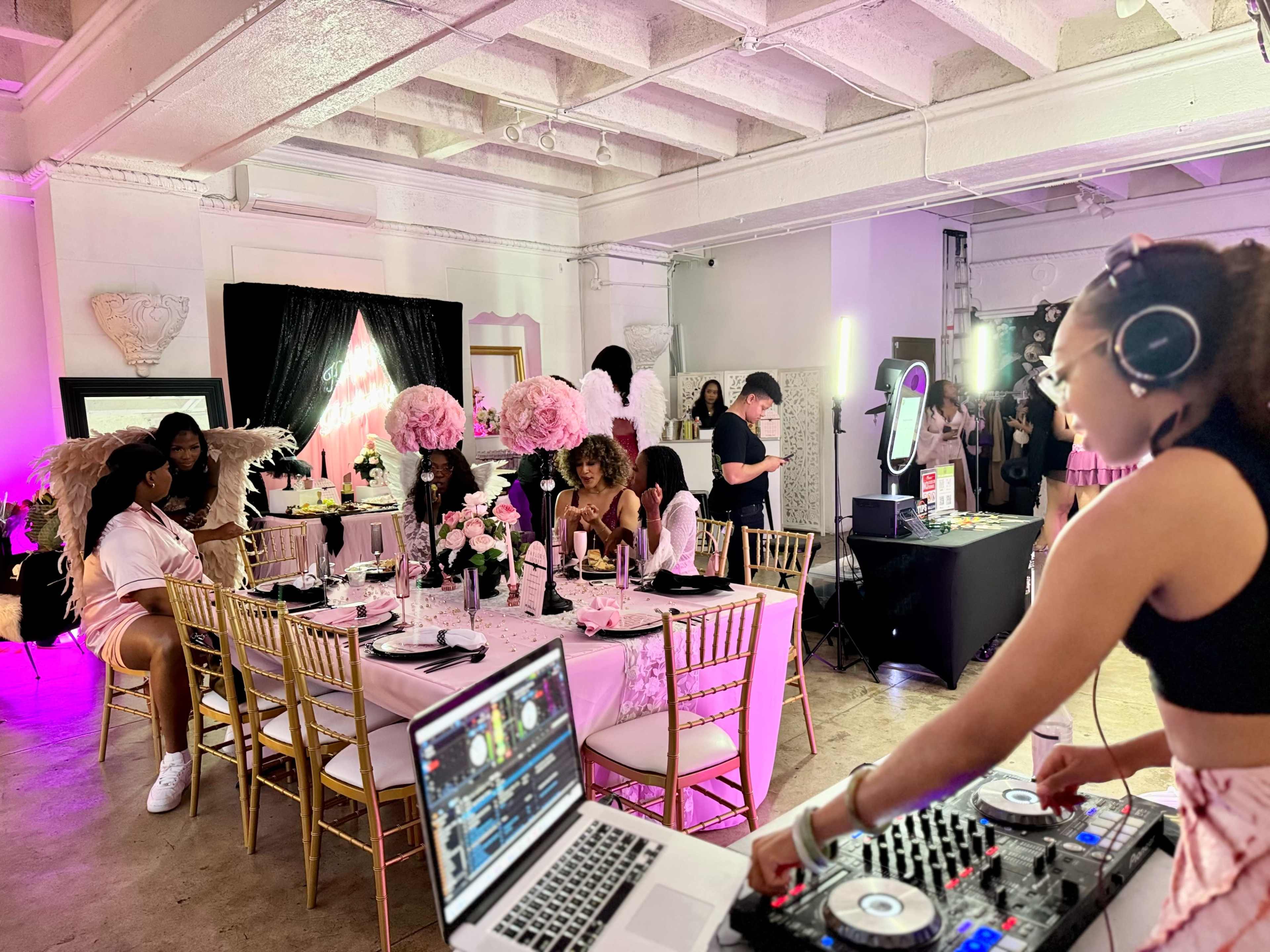 The scene shows a vibrant indoor party with guests seated at elegantly decorated tables, a DJ mixing music in the foreground, and pink and gold decorations throughout the space.