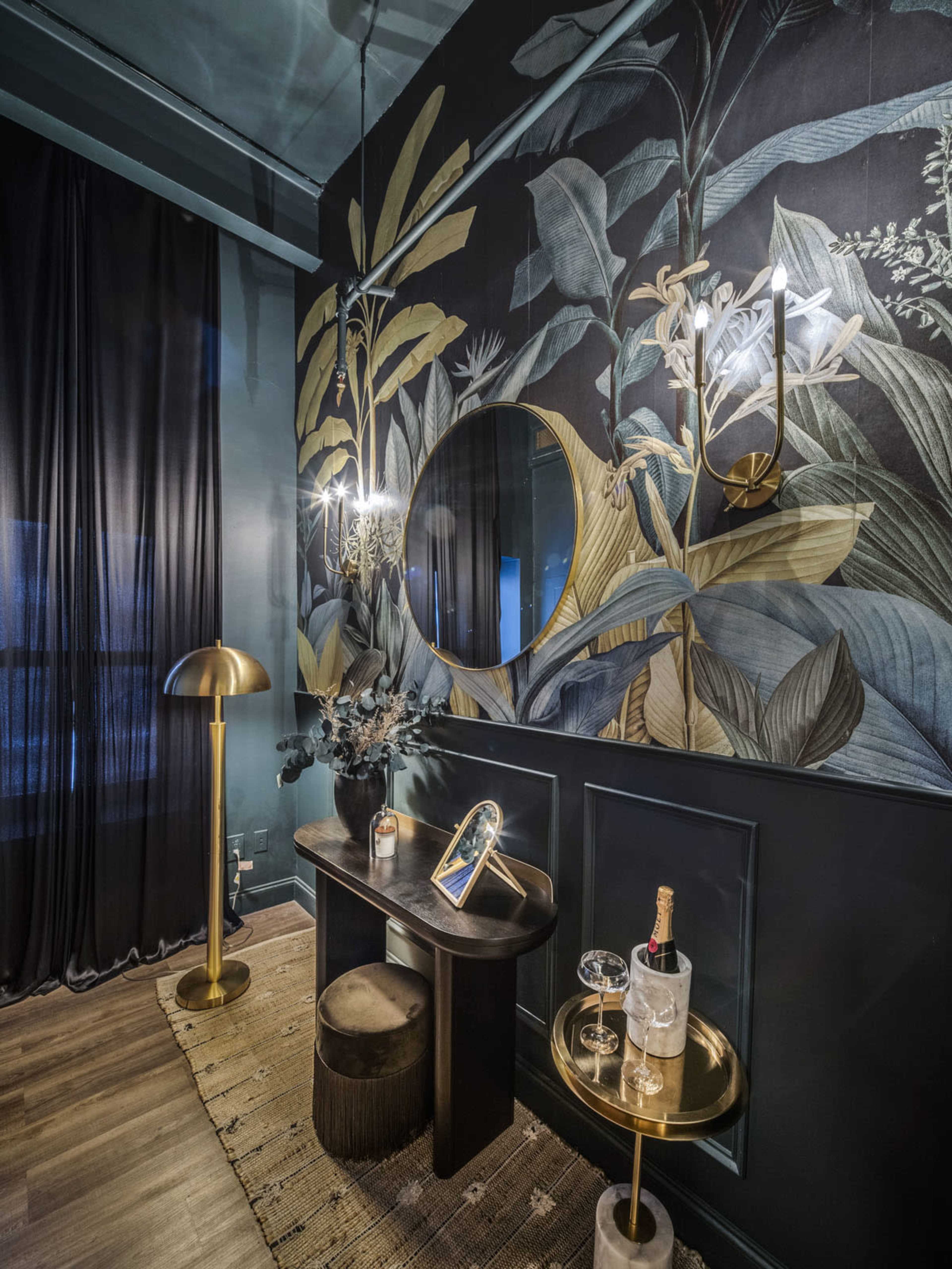 The image depicts a stylish room interior featuring a dark leaf-patterned wall, a round mirror, a small table with drinks, and a floor lamp.