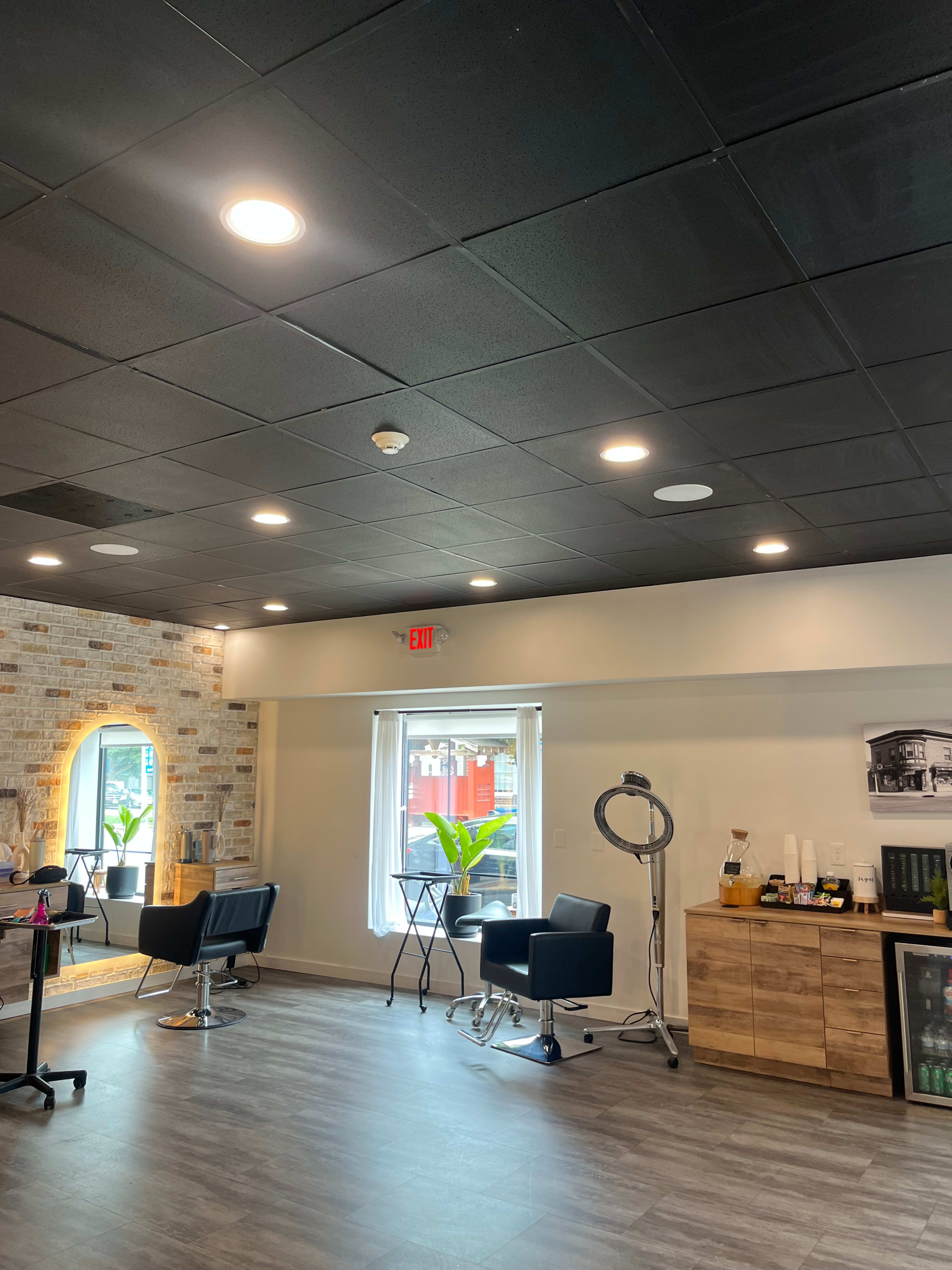 The image shows a modern salon interior with black ceiling tiles, wooden furniture, and a large window letting in natural light.