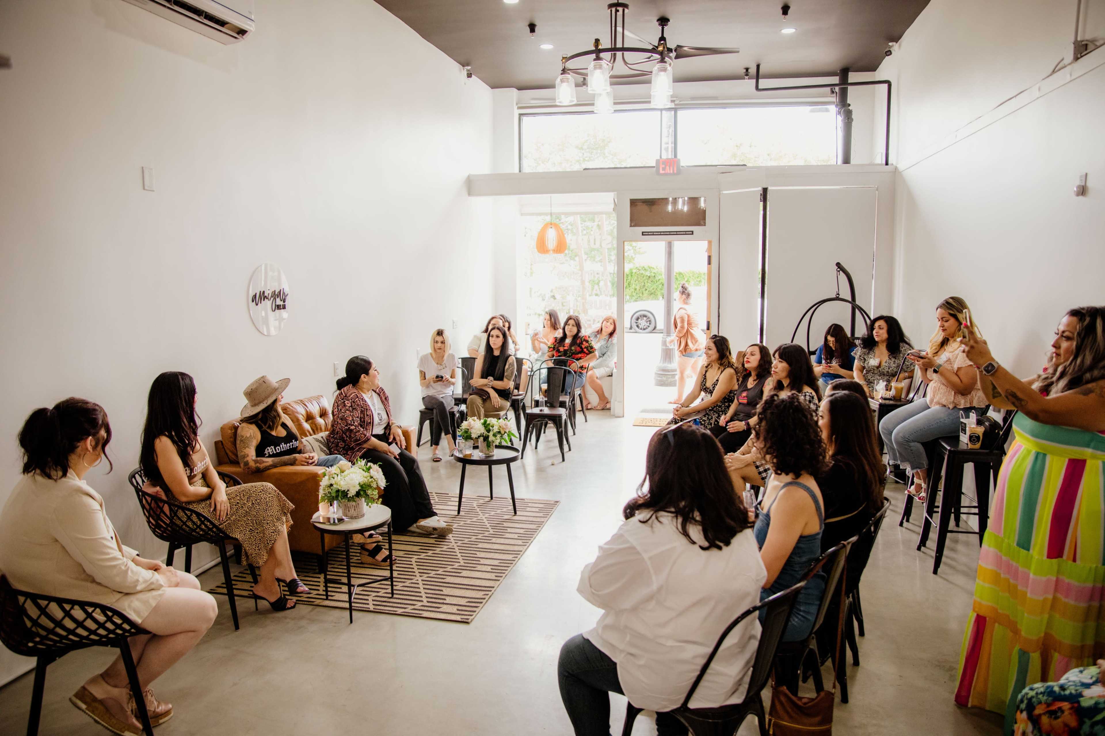 A group of women are seated in a modern, sunlit venue for a gathering, with some engaging in conversation while others listen.