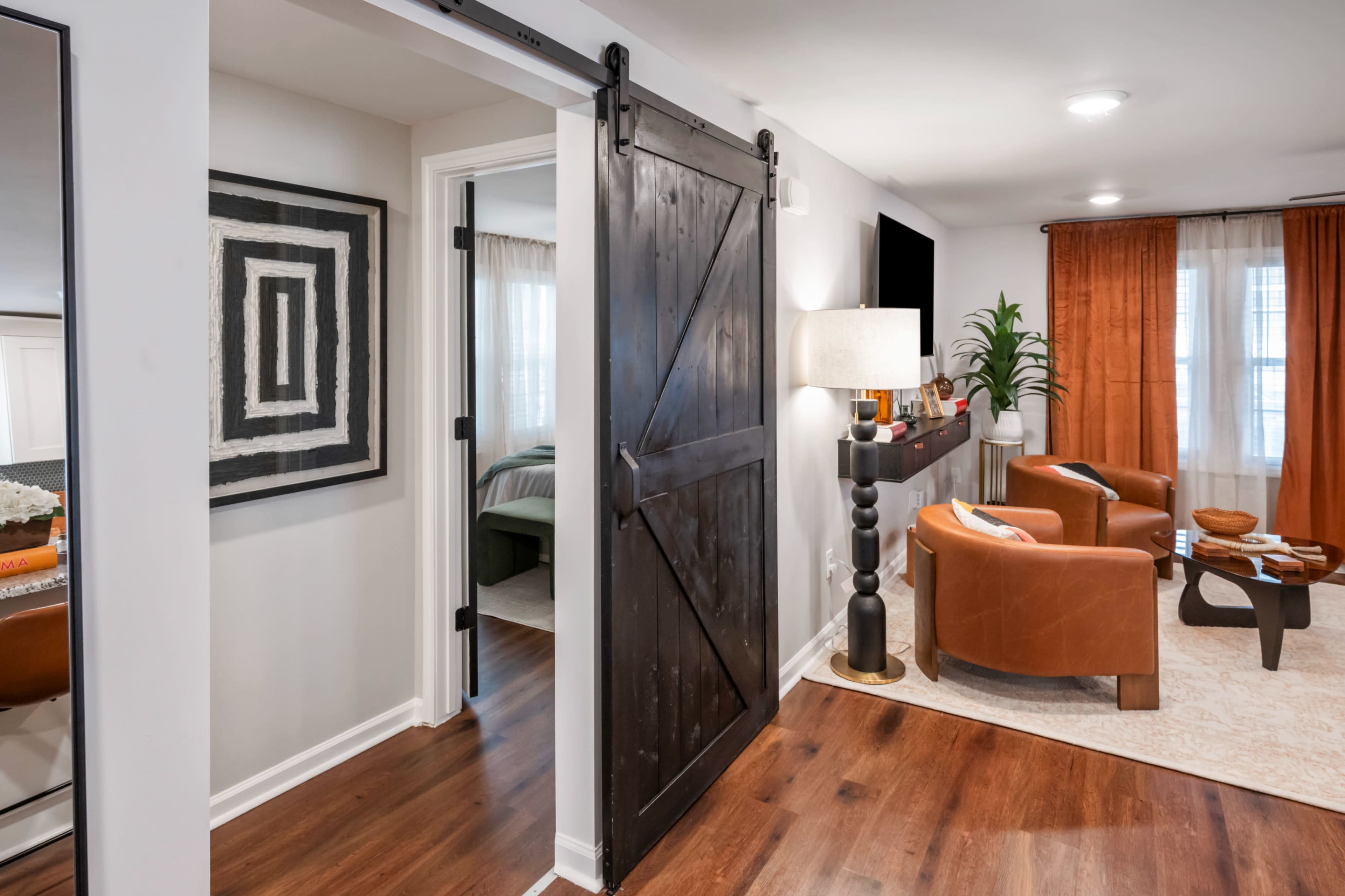 The image shows a modern living space with a sliding barn door, two brown chairs, a lamp, and an orange curtain, along with a glimpse of a bedroom through the doorway.