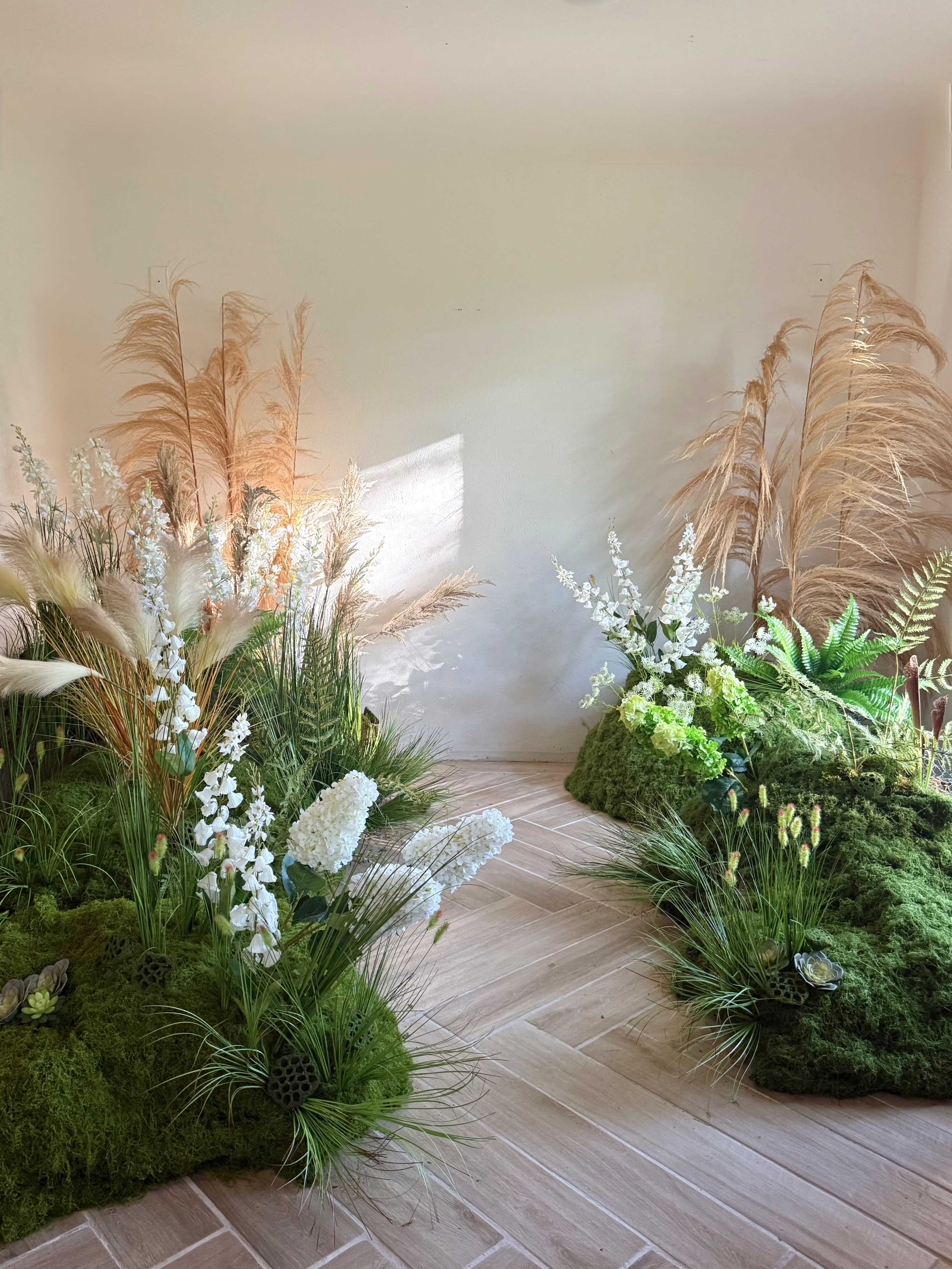 The image shows a decorated indoor space with green mossy arrangements and various artificial flowers and plants, creating a lush, natural aesthetic.