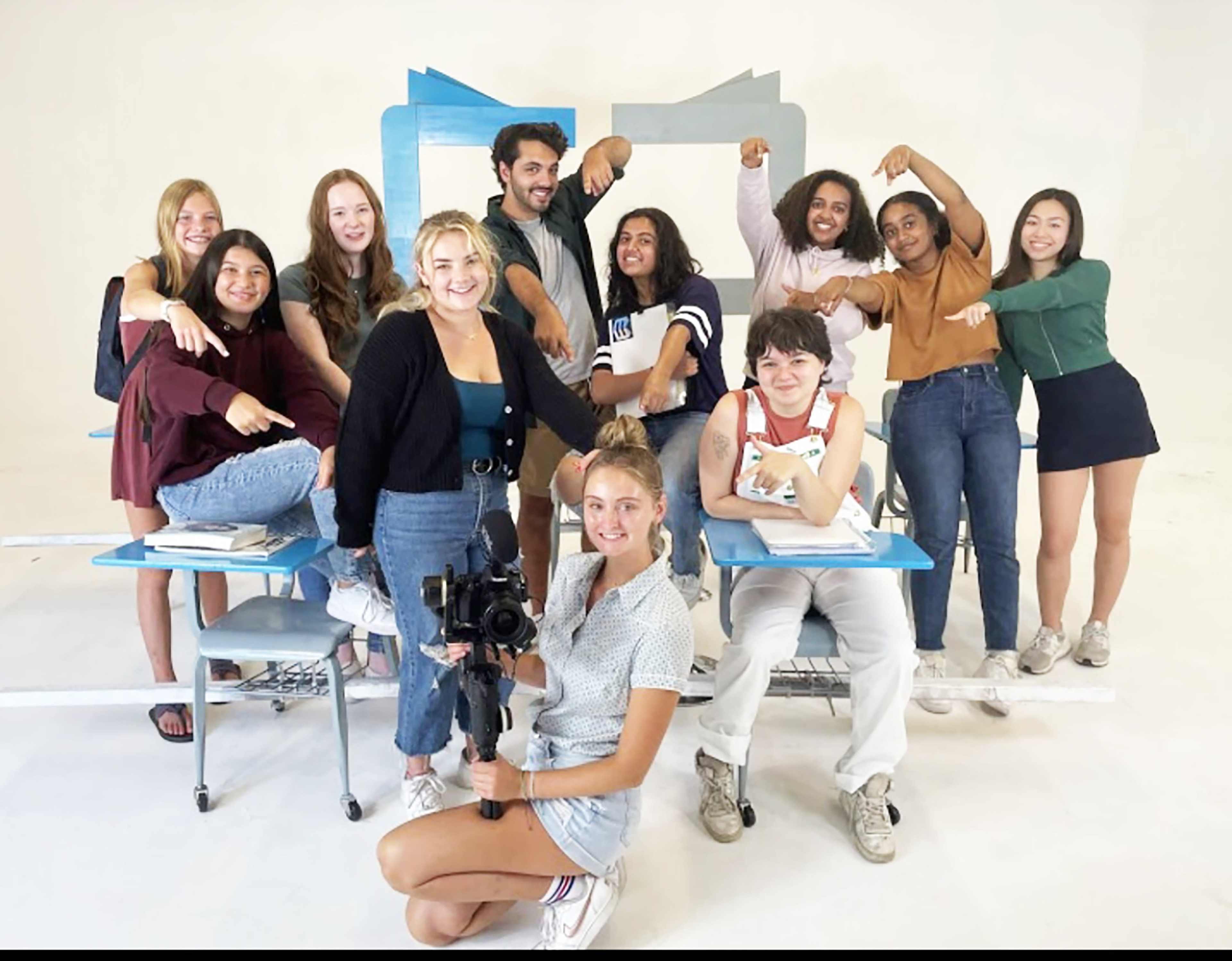 A group of twelve teenagers poses for a photo in a studio setting with desks and a backdrop.