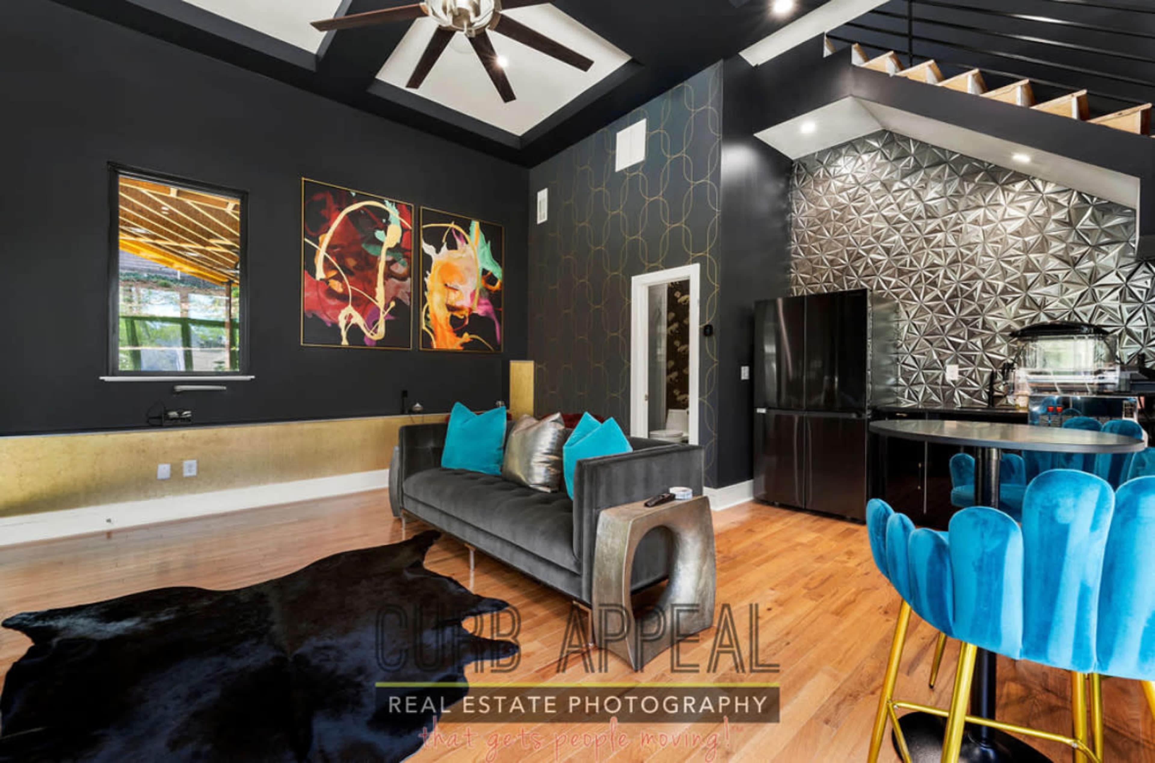 The image depicts a modern interior featuring a living area with a dark sofa, turquoise accent pillows, vibrant artwork on the walls, and a kitchen with a geometric backsplash.