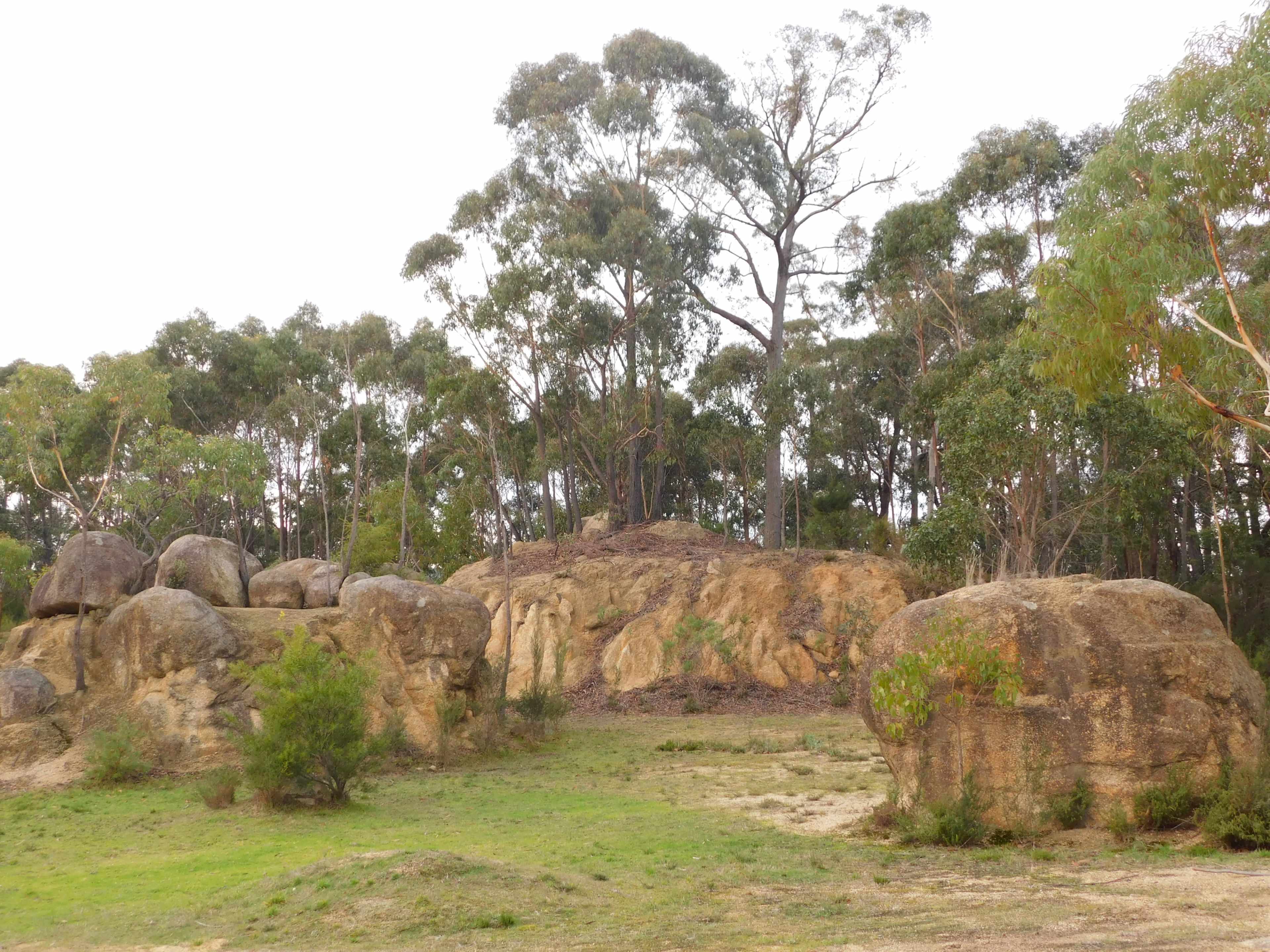 A cluster of large boulders is situated beside a hill adorned with trees amidst a grassy area.