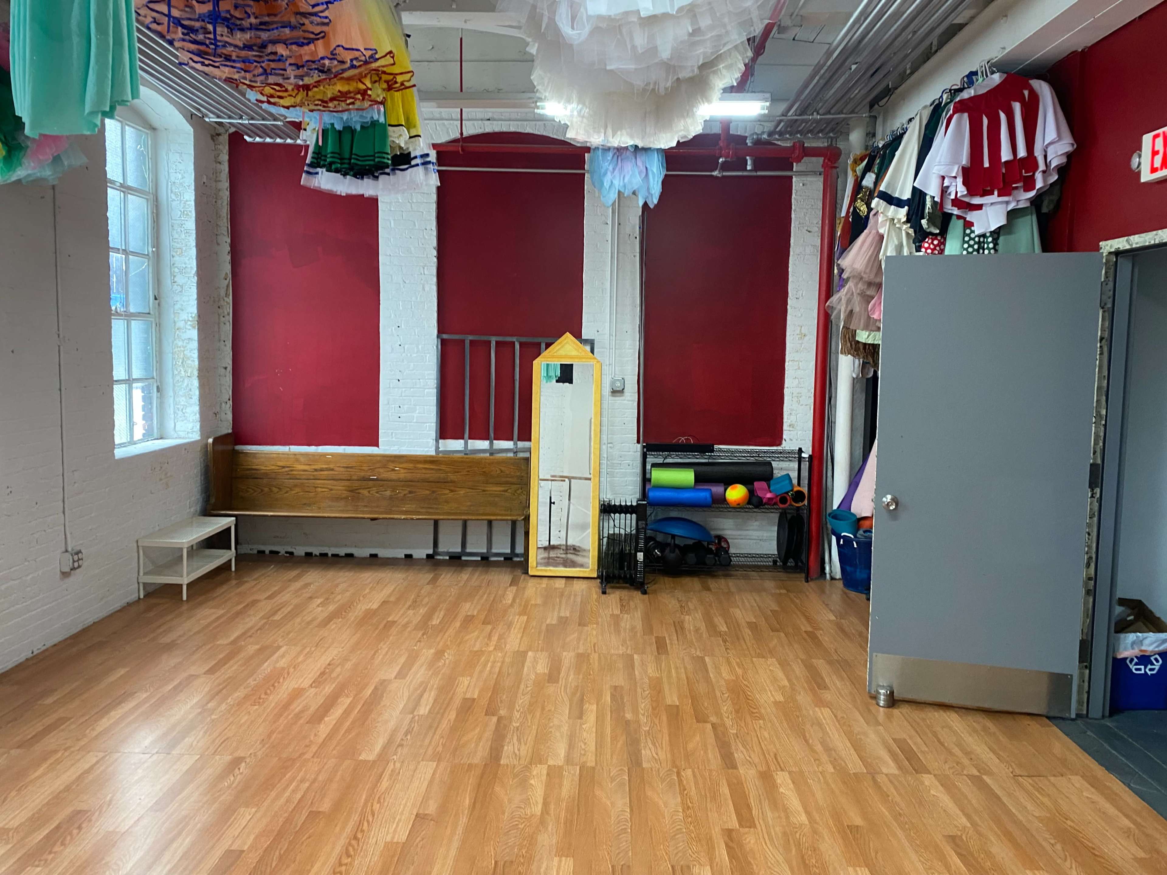 A spacious room with wooden flooring, white brick walls, and a mirror alongside a bench, featuring colorful fabric and exercise equipment stored in the corner.