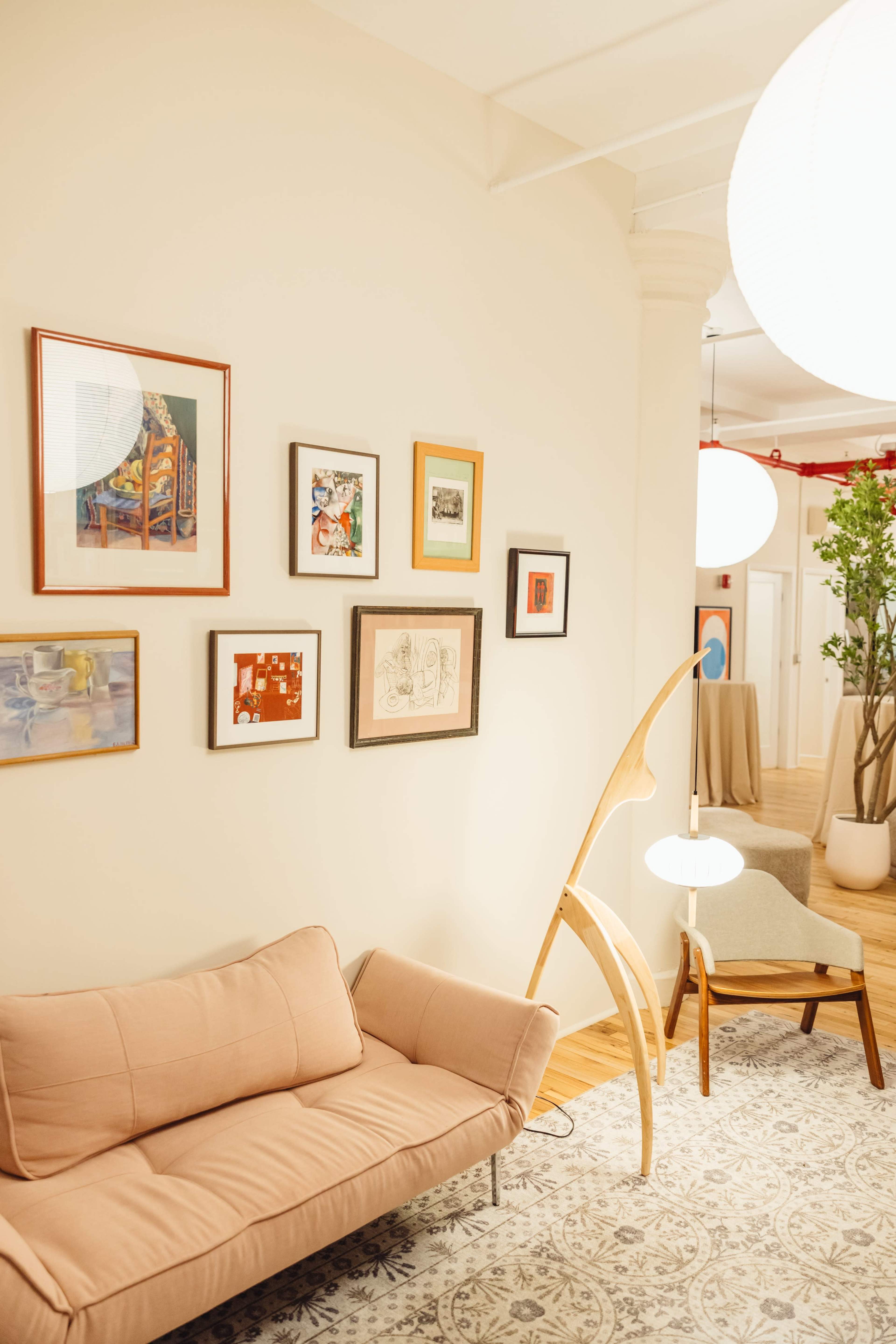 The image shows a cozy interior with a pink sofa, framed artworks on the wall, a sculptural floor lamp, and a decorative plant in a large pot.