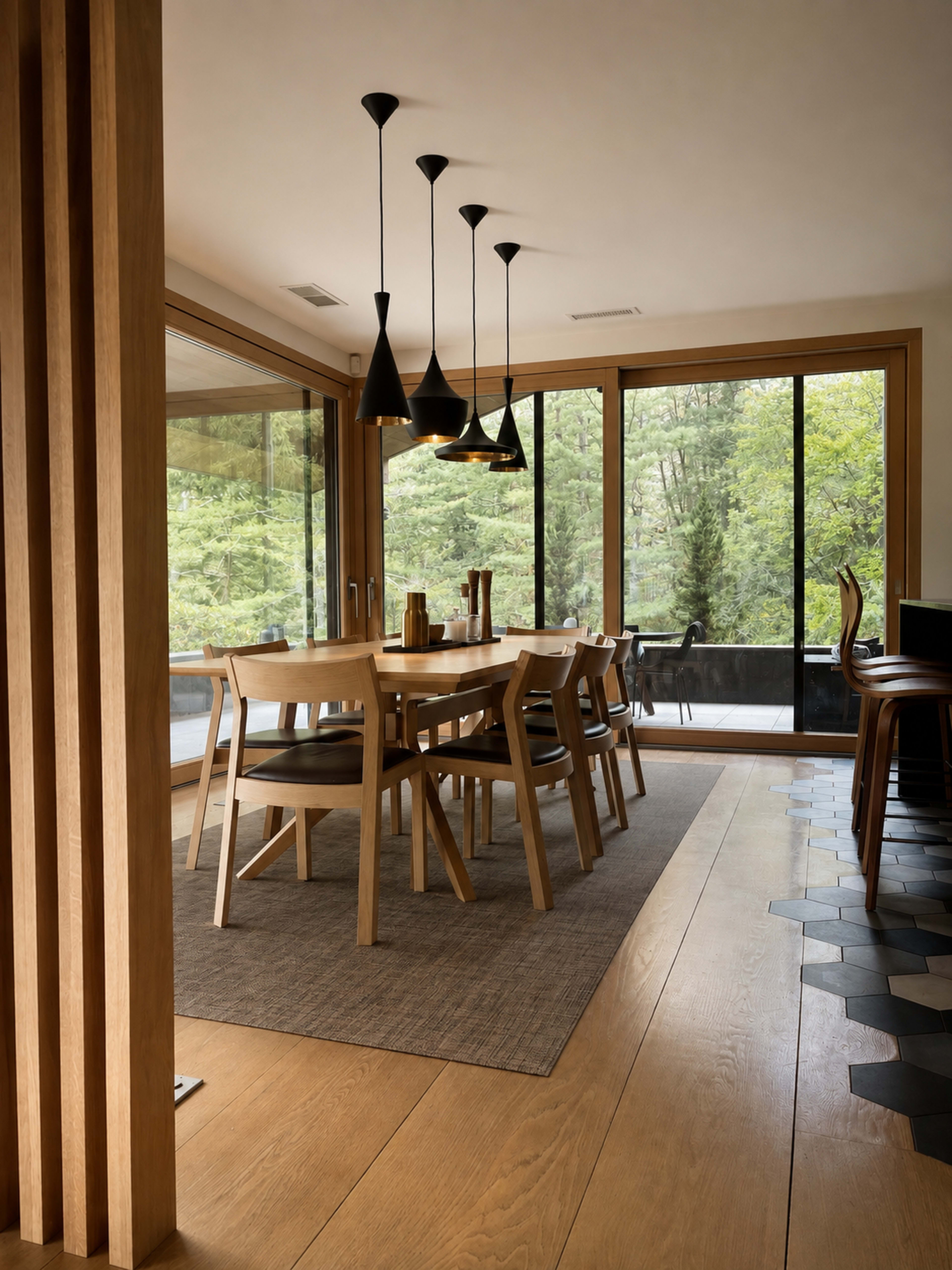 The image shows a modern dining area featuring a wooden table surrounded by chairs, with large windows overlooking greenery and pendant lights hanging from the ceiling.