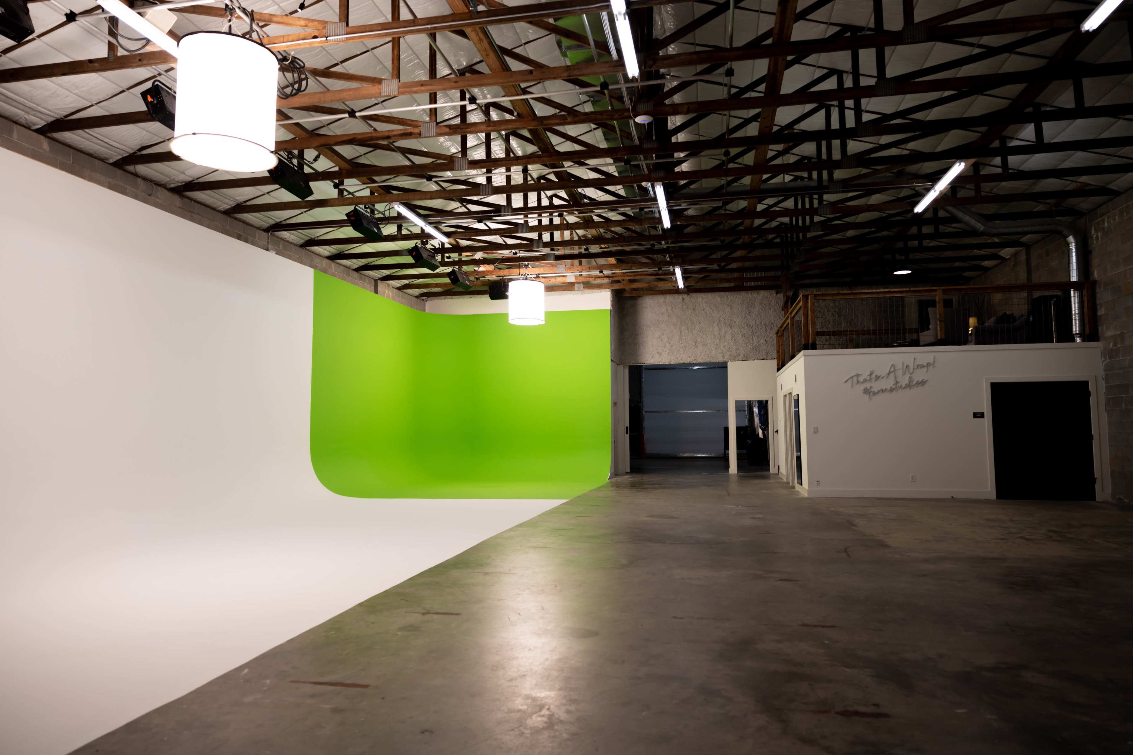 The image shows an industrial studio space featuring a bright green screen on one side and a white backdrop on the other, with exposed wooden beams and overhead lighting.