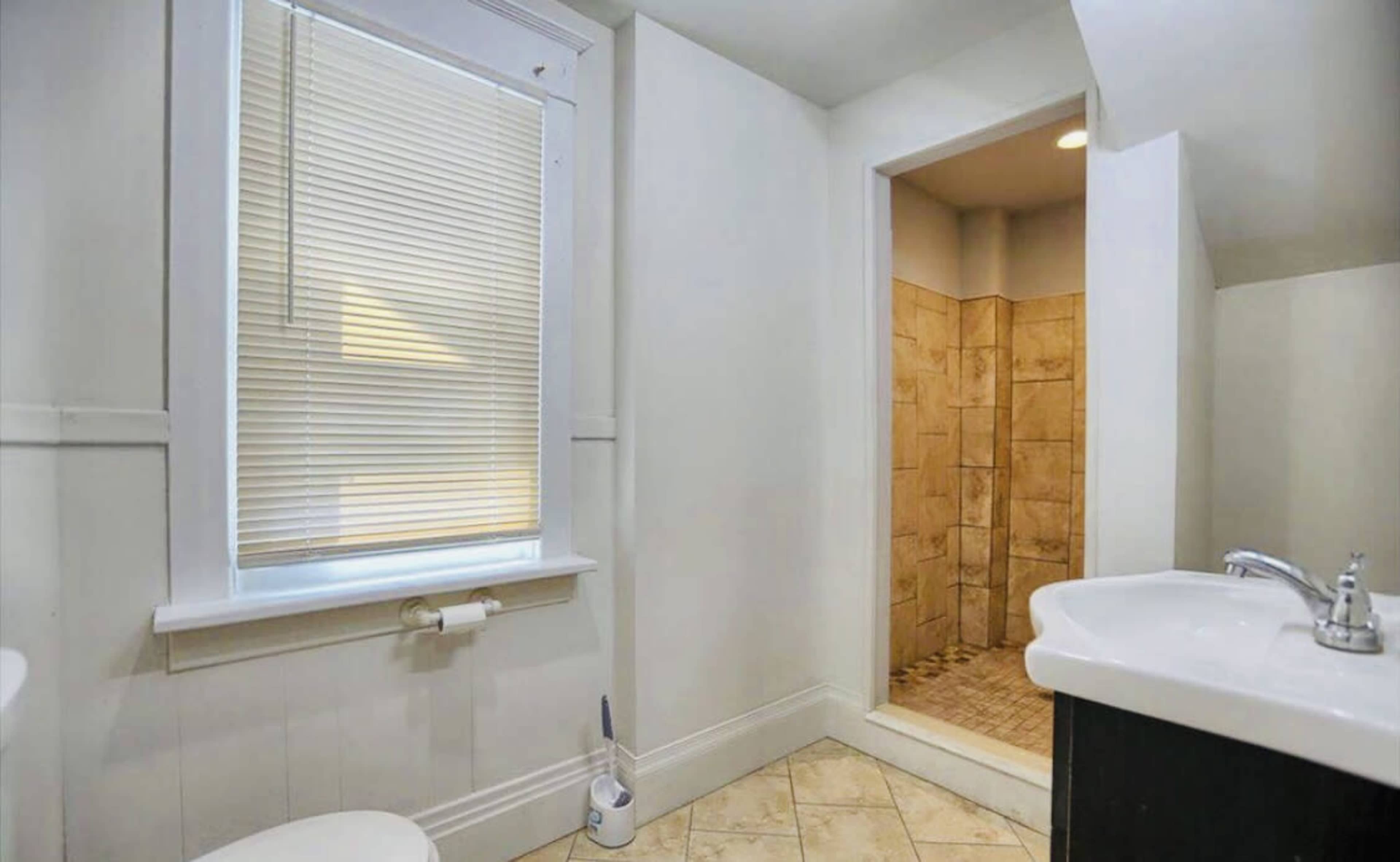 A bathroom with a toilet, sink, and a separate shower area, featuring tiled walls and a window with blinds.