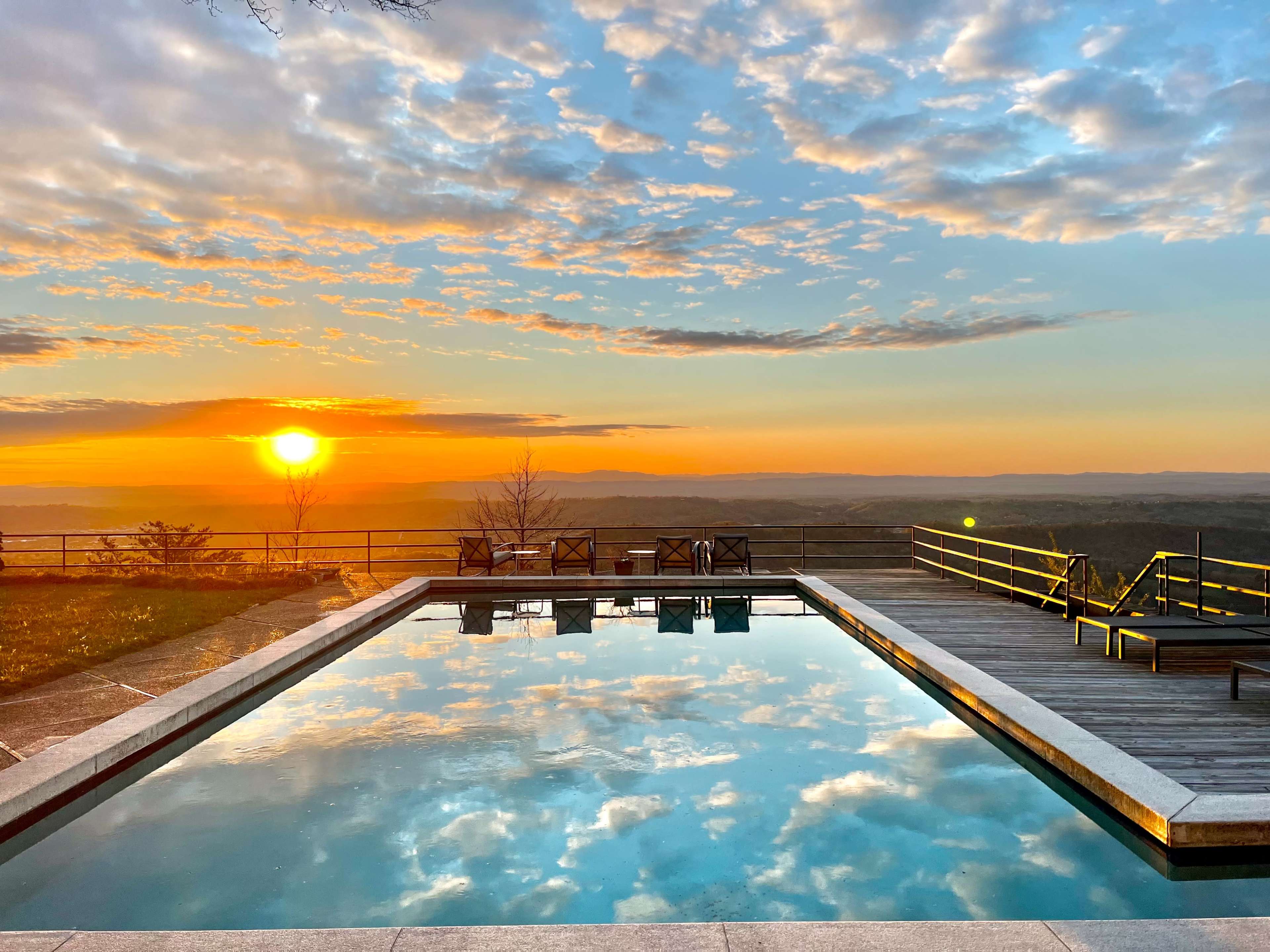 A pool reflecting a sunset with clouds, framed by a wooden deck and empty chairs overlooking a valley.