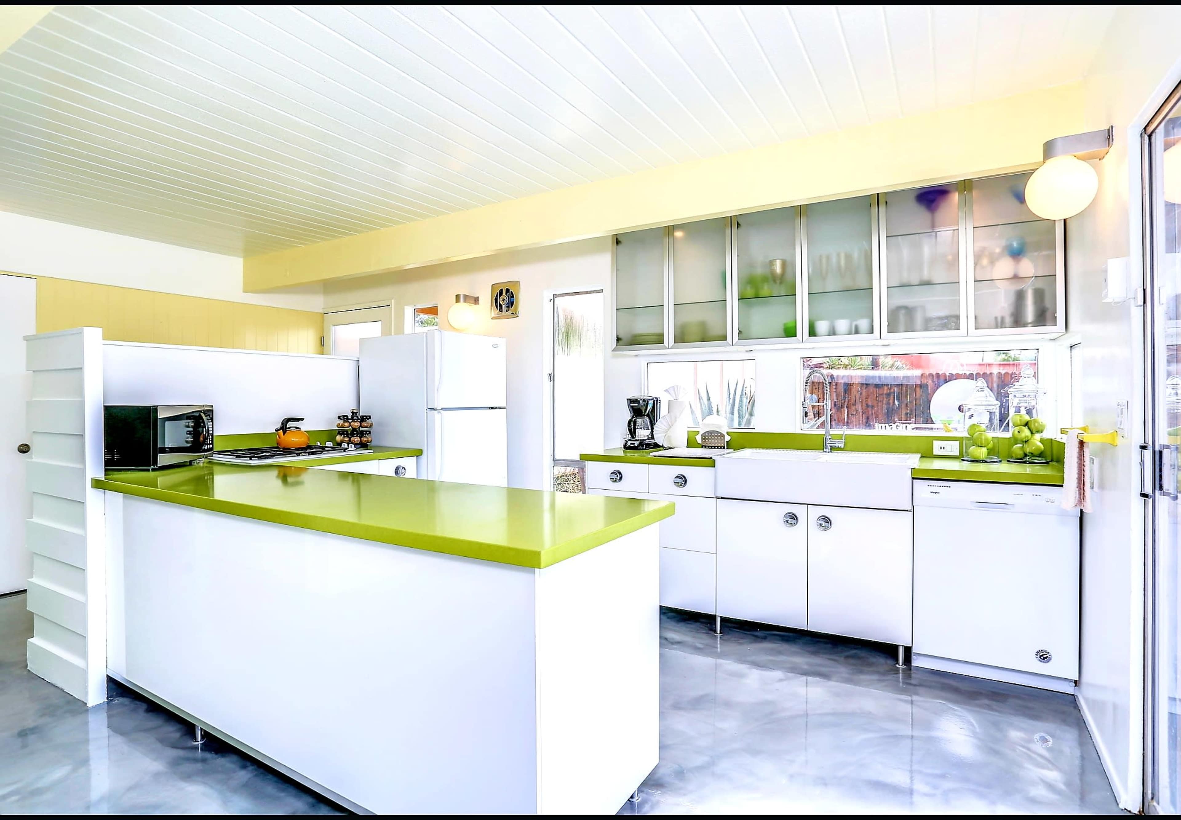 The image shows a modern kitchen with white cabinetry and green countertops, featuring a refrigerator, microwave, and coffee maker on a large island.