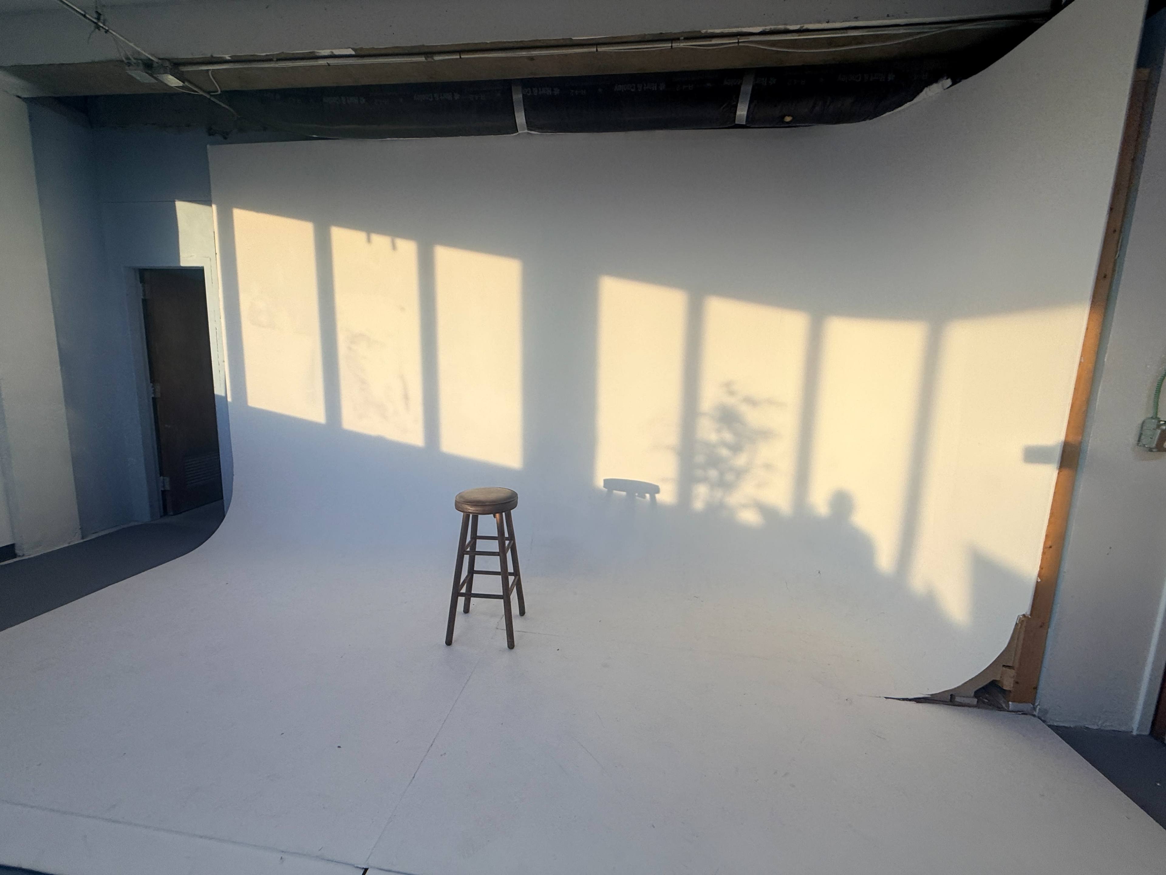 A wooden stool sits on a white backdrop under natural light, with shadows cast on the wall from nearby windows.