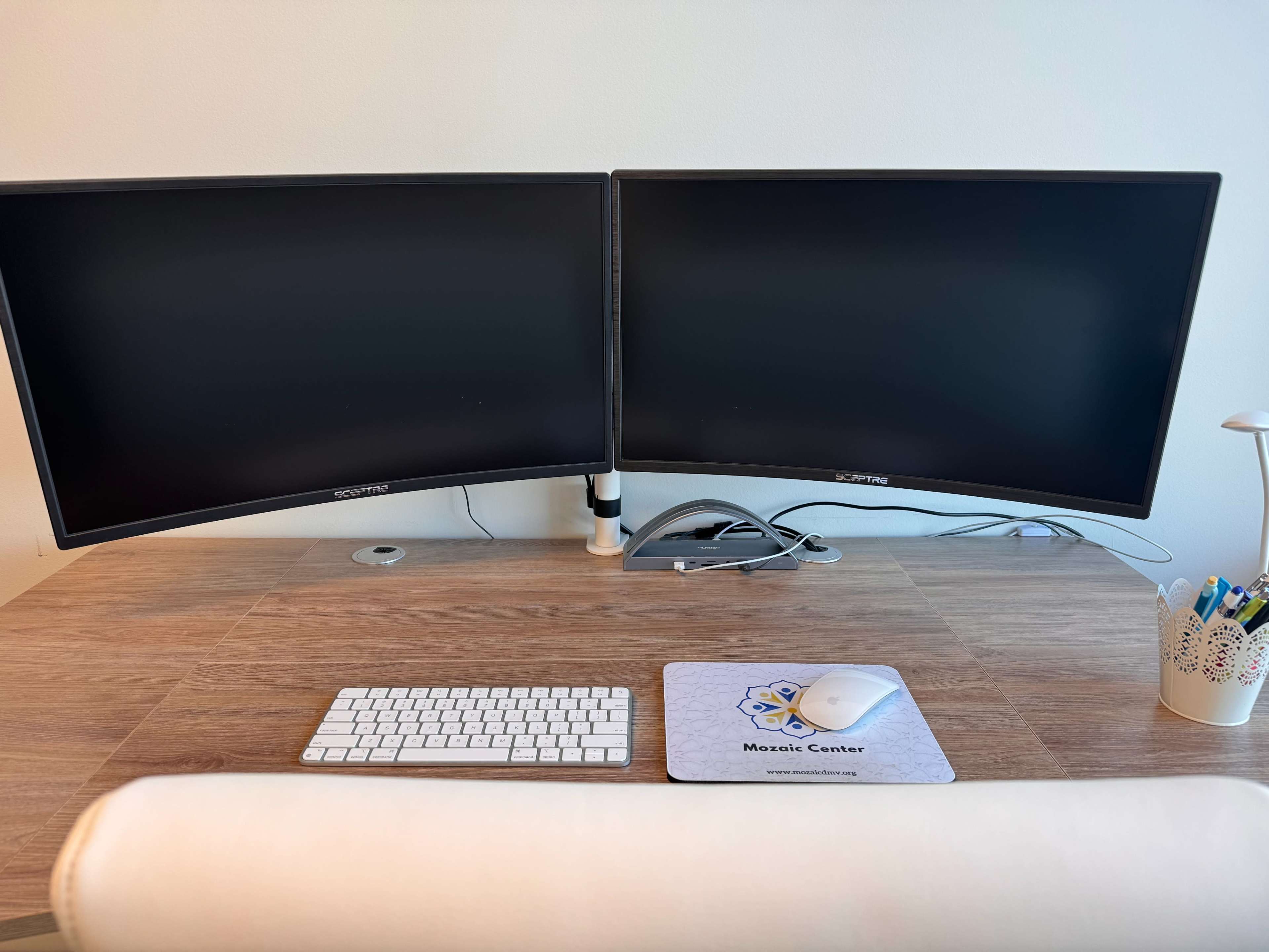 A desk with two curved computer monitors, a white keyboard, and a mouse on a mouse pad, accompanied by a pen holder and a lamp.