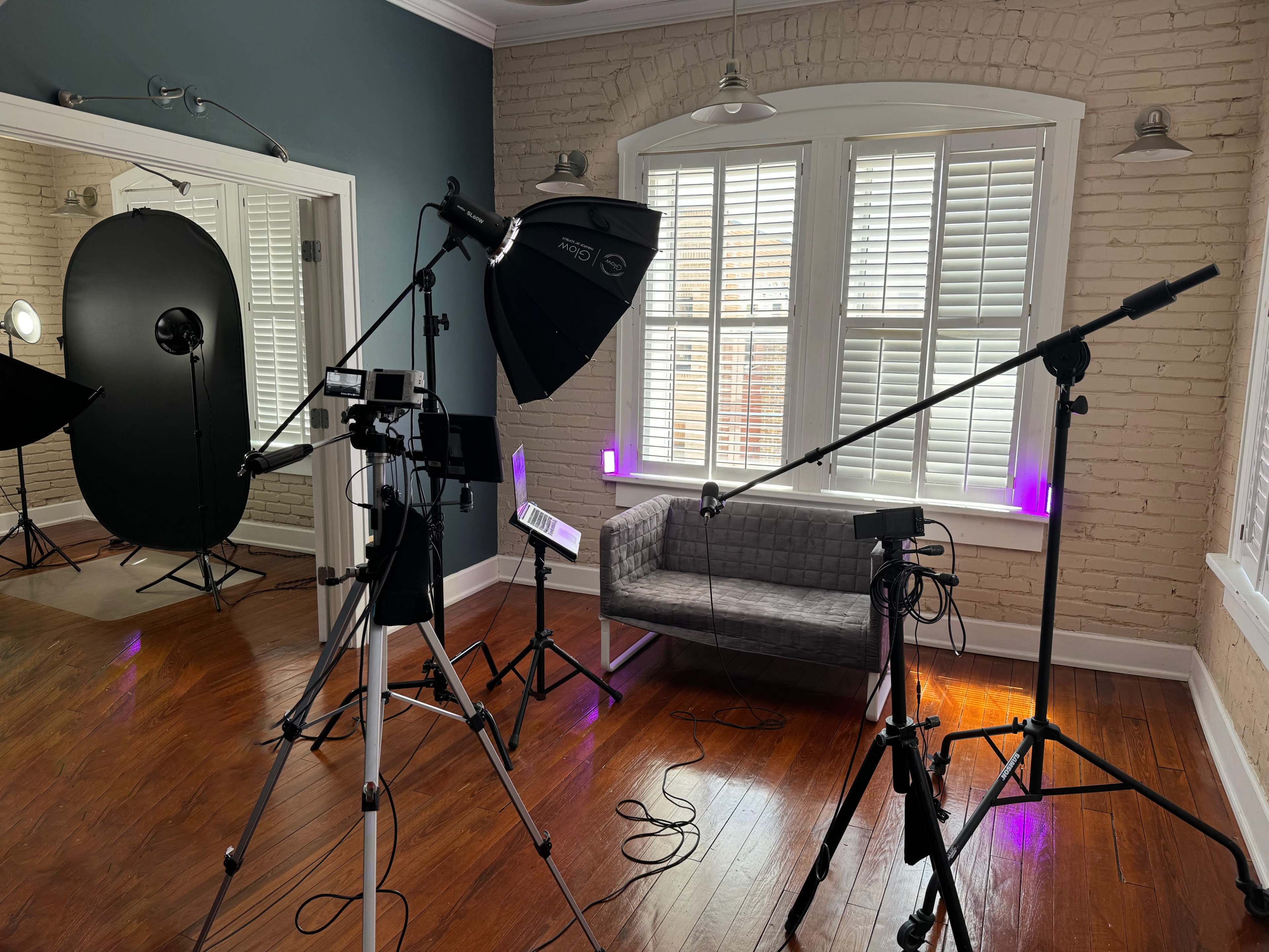 A photography studio setup with multiple tripods, lighting equipment, and a gray couch against a brick wall.
