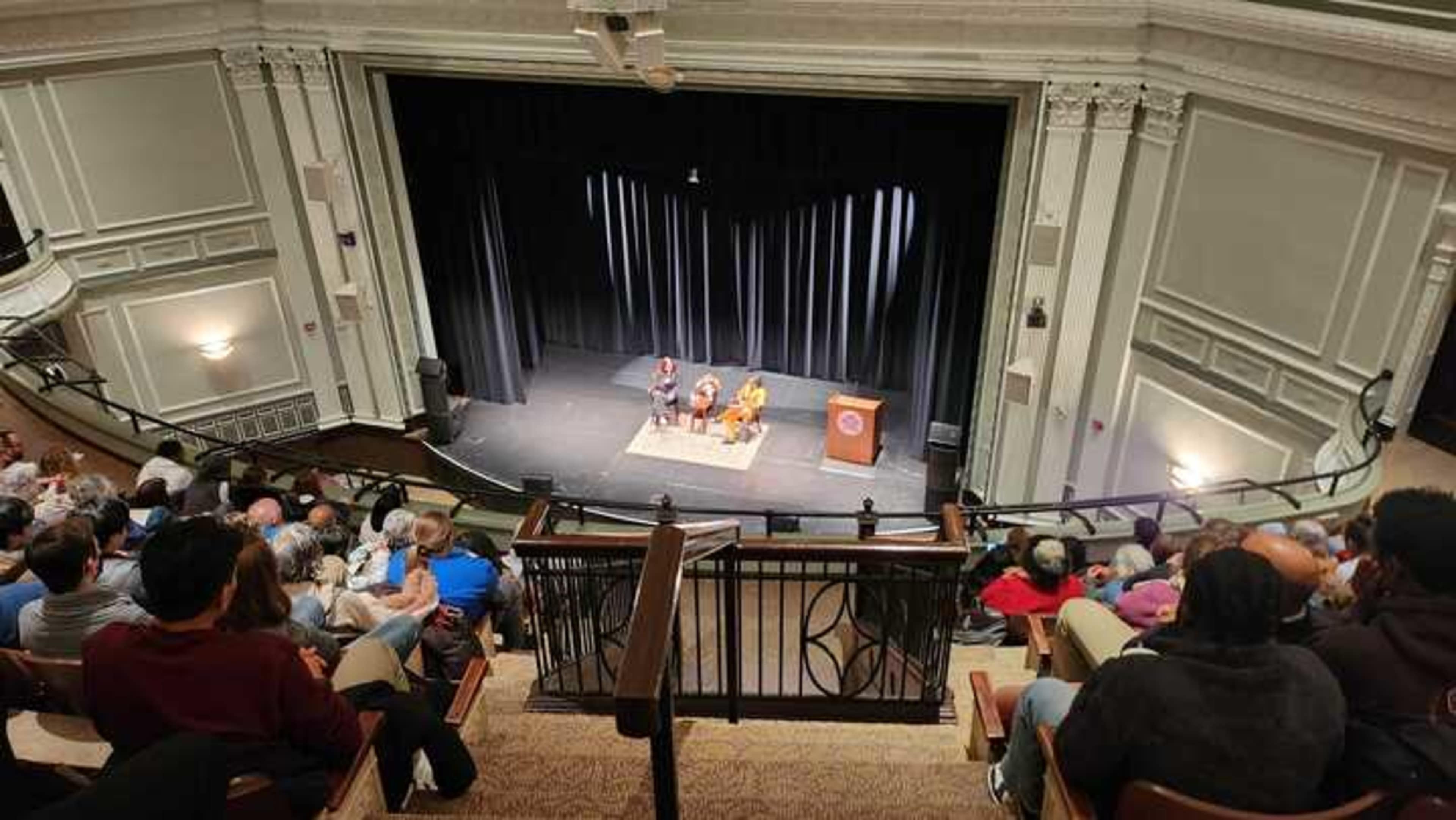 A speaker event is taking place on a stage in an auditorium, with an audience seated in rows.