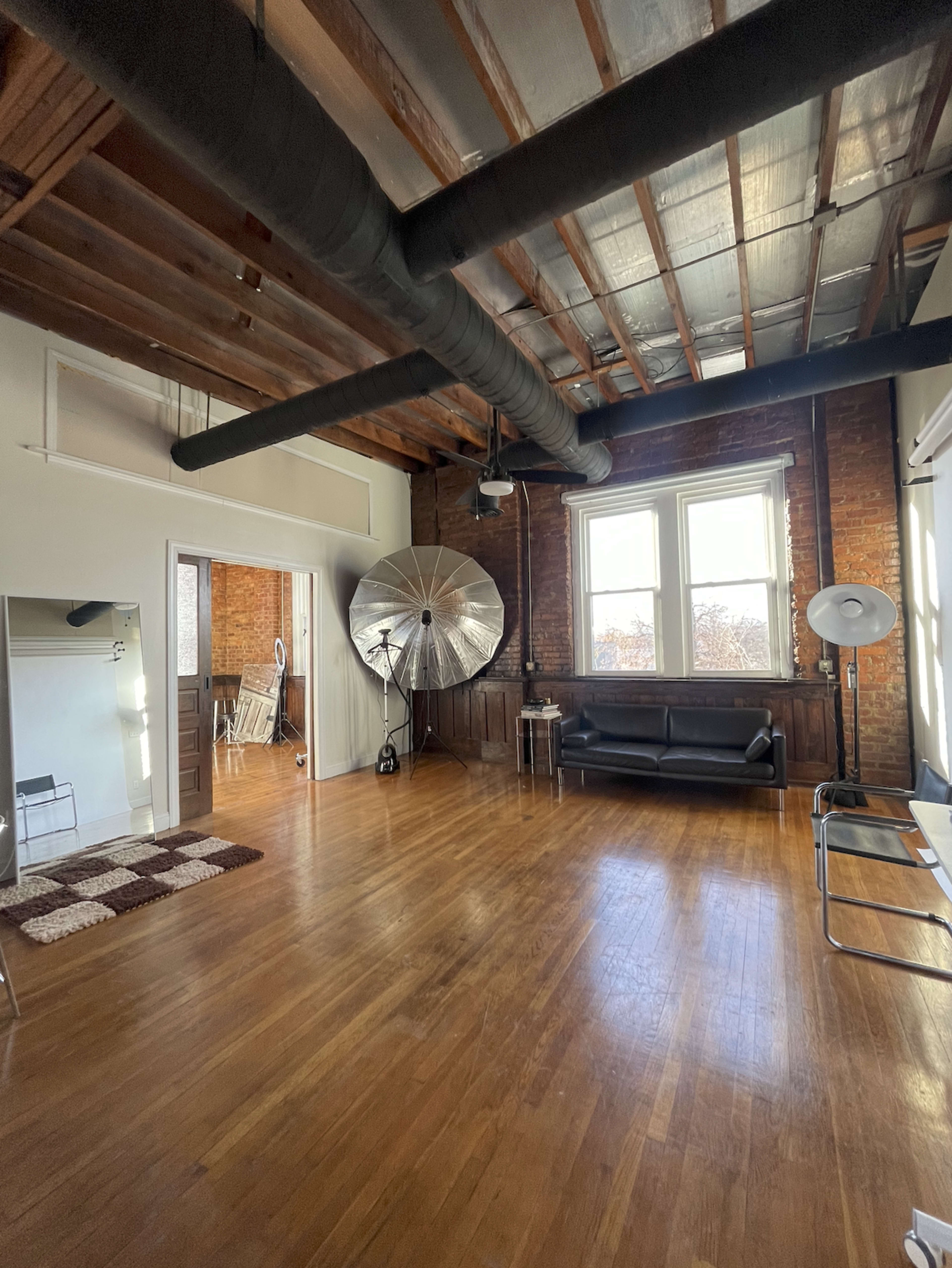 A spacious room with wooden floors, exposed brick walls, and large windows, featuring a black sofa, a large silver reflector, and a few pieces of minimalist furniture.
